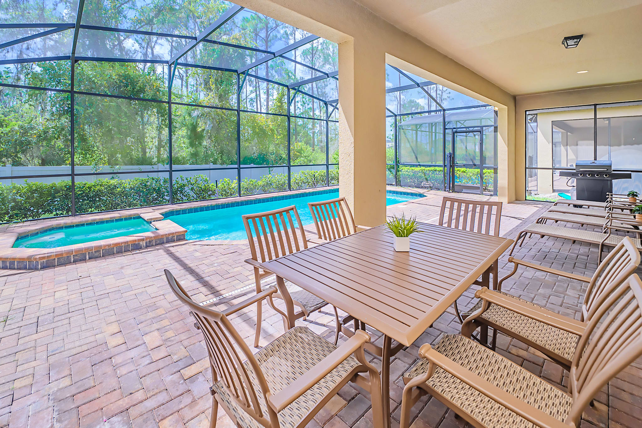 Covered outdoor patio with a wooden dining table and six chairs, overlooking a screened-in private pool and hot tub area, with lush greenery visible beyond the screen.