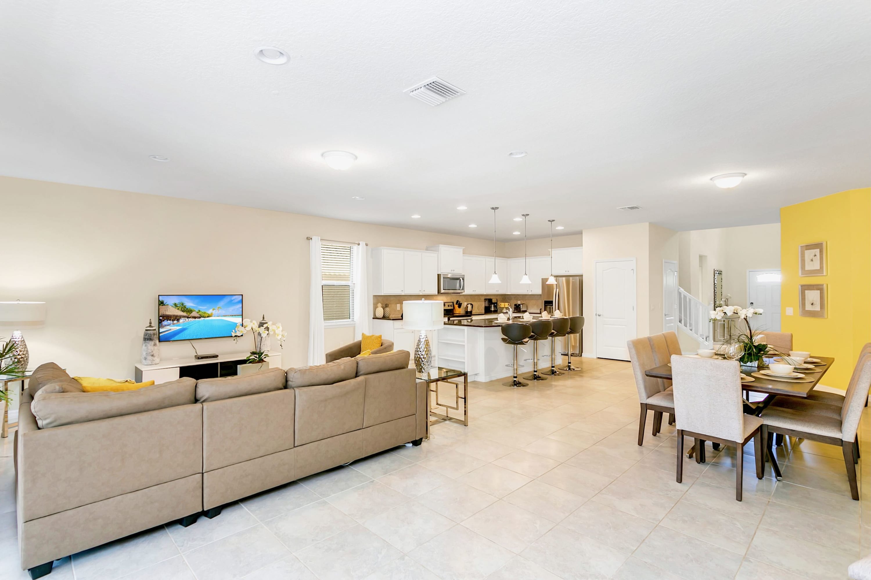 Bright open-concept living space with a large L-shaped sectional sofa facing a TV, a dining area with a wooden table and upholstered chairs, and a modern kitchen island with bar stools. The room features light tile flooring and neutral walls.