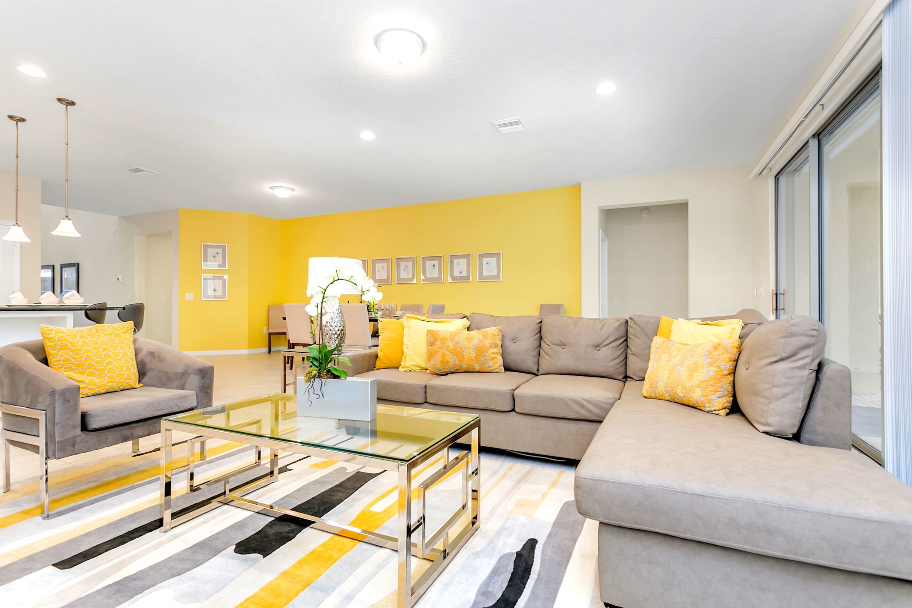 Bright living room with a large L-shaped sectional sofa and an armchair in gray, accented with yellow pillows, a glass coffee table, and a striped rug, leading to a dining area with a vibrant yellow accent wall.