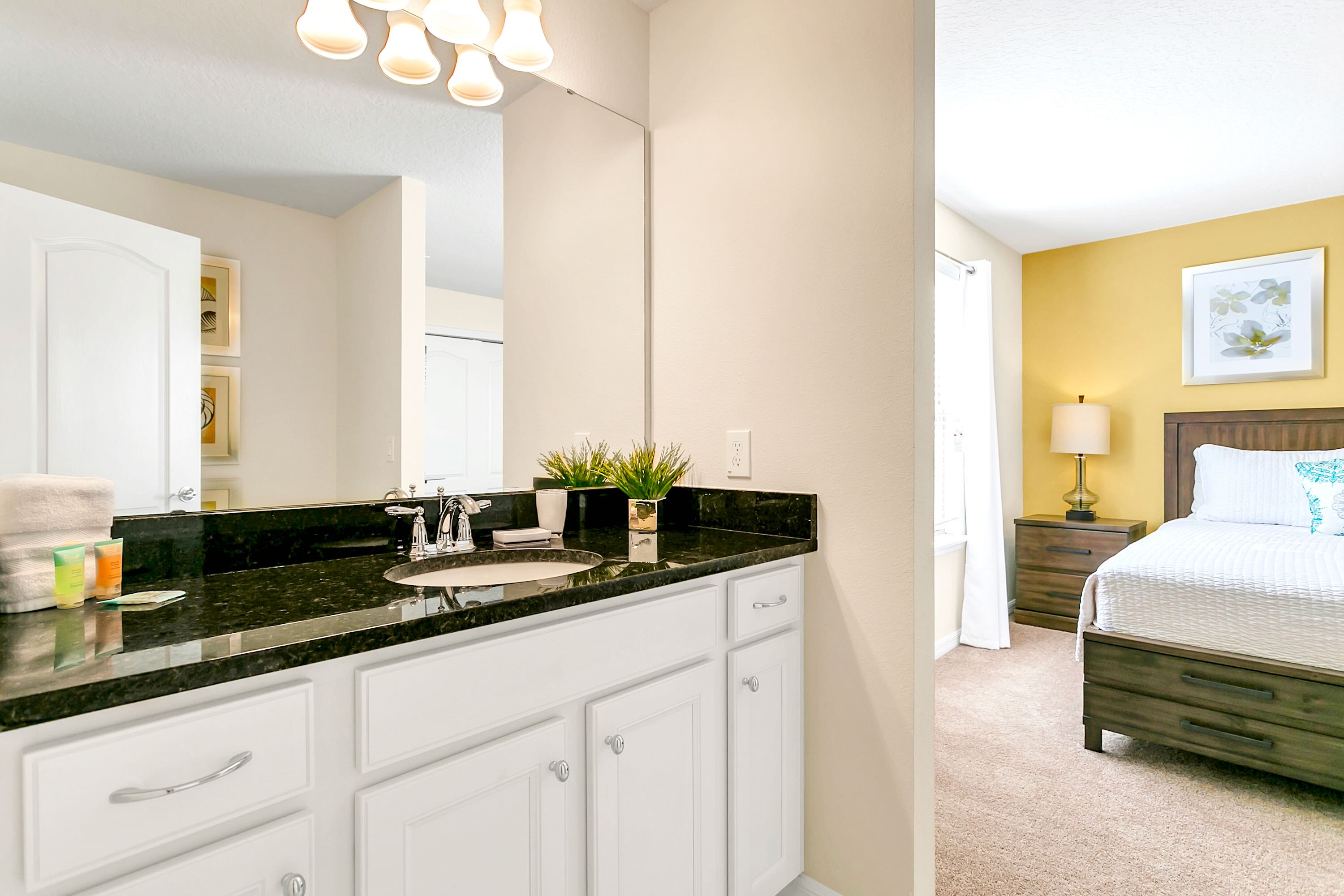 This bathroom features a dark granite countertop with an undermount sink, white cabinetry, and a large mirror reflecting a portion of a bedroom with a yellow accent wall and a bed.