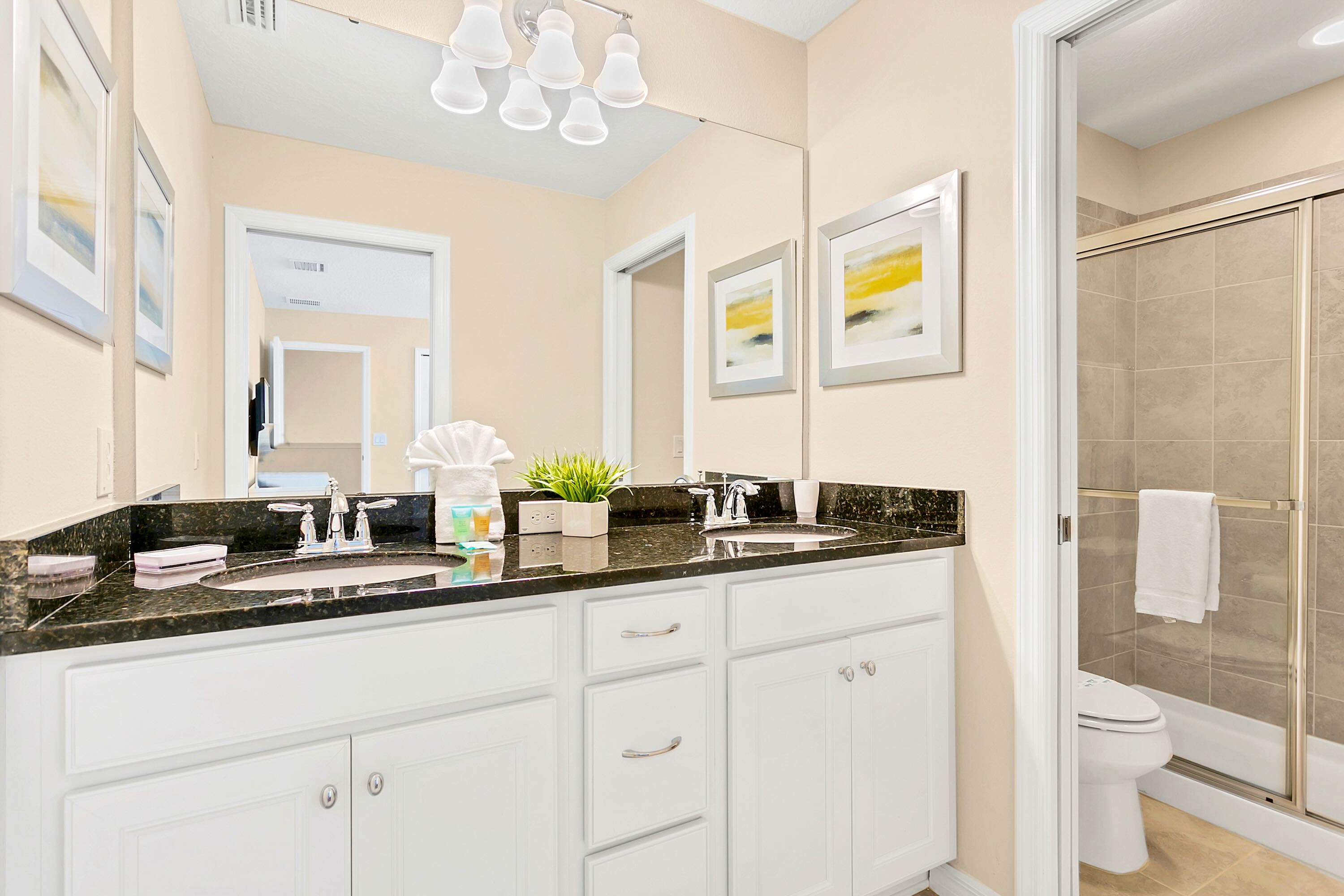 A bright bathroom with a double vanity, dark granite countertops, white cabinets, and a large mirror reflecting a bedroom with a yellow accent wall. The shower-tub combo with a sliding glass door and toilet are visible to the right.