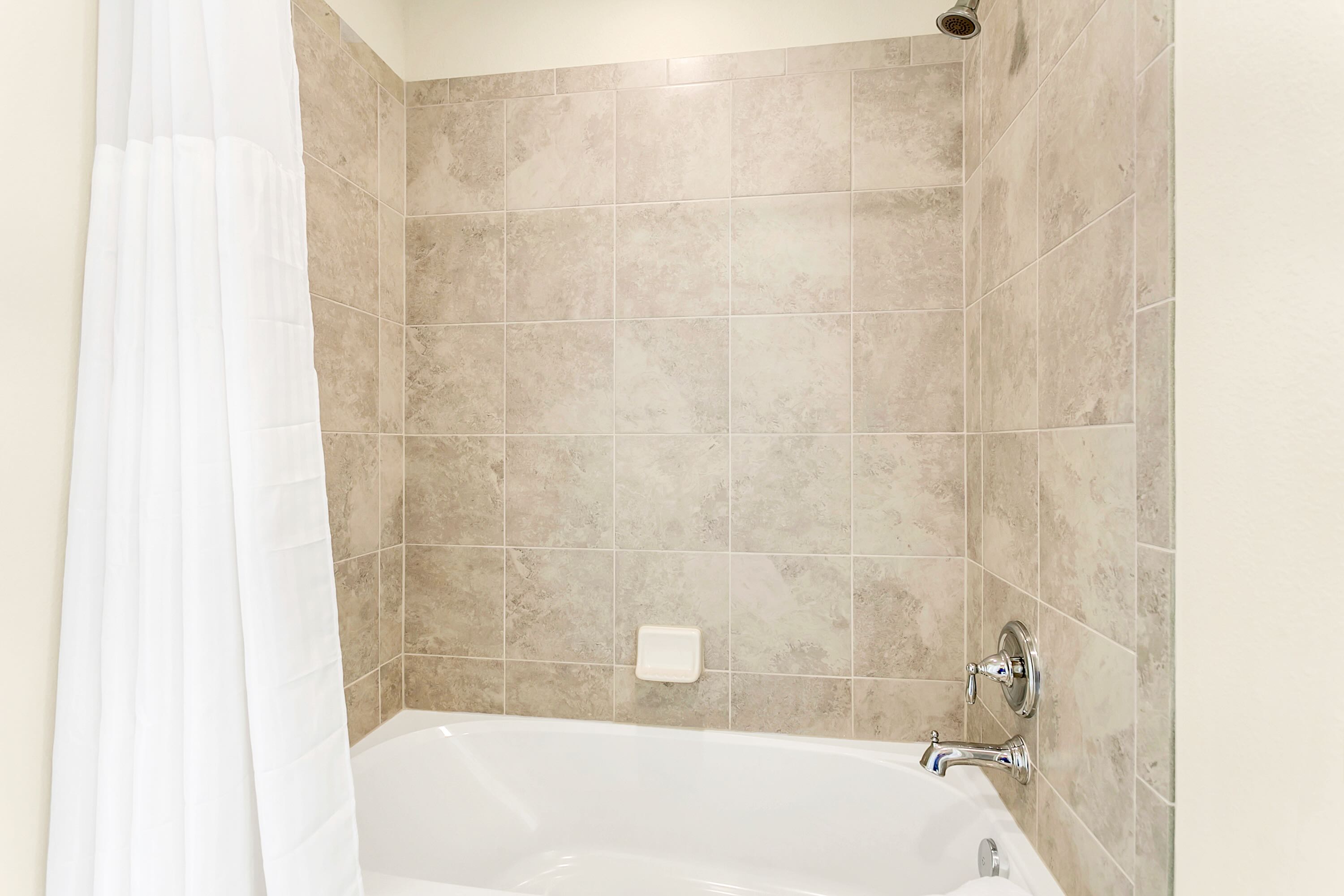 A clean white bathtub is set against light beige tiled walls, featuring a chrome showerhead and faucet, and a white shower curtain on the left.