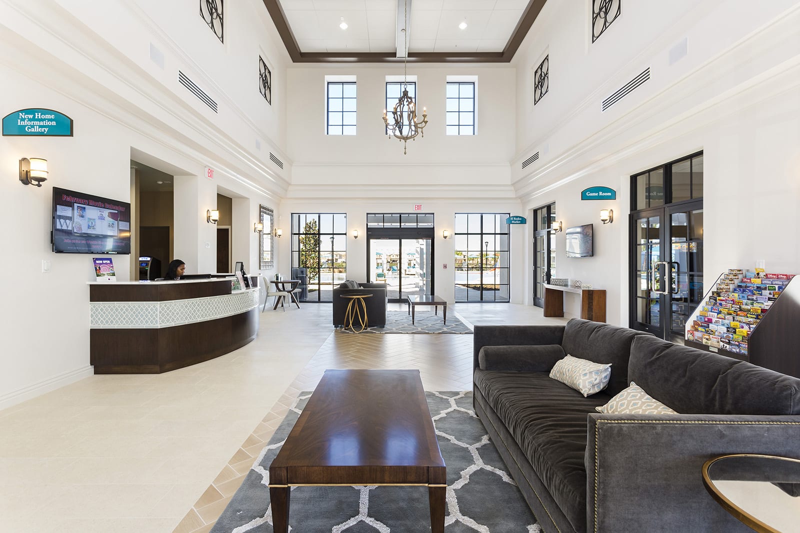 A spacious and elegant clubhouse lobby featuring a reception desk with a patterned front, high ceilings with a chandelier, and a comfortable seating area with a dark grey sofa and a wooden coffee table on a patterned rug. Large windows and glass doors provide natural light.