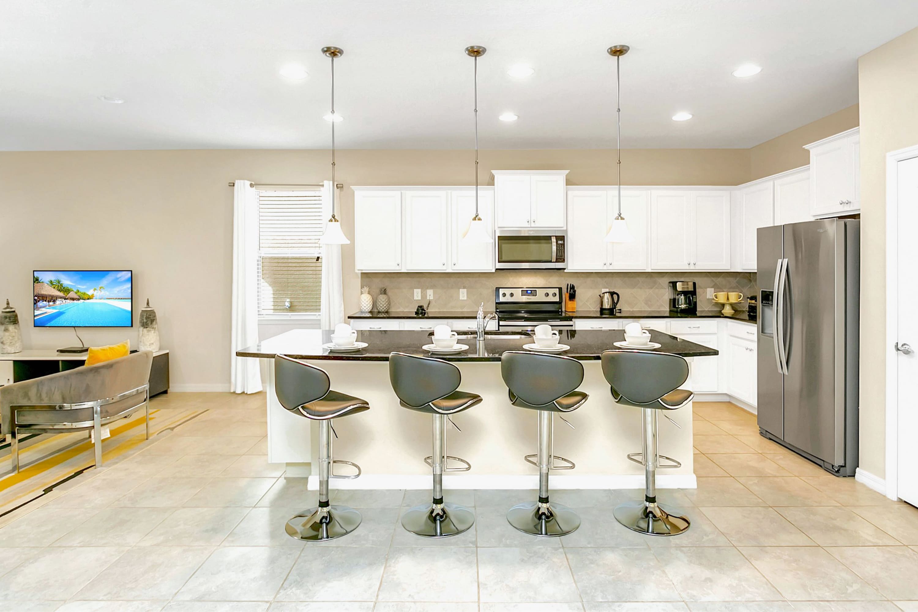 Modern kitchen island with four dark upholstered bar stools, facing an open living area with a television on a console. The kitchen features white cabinetry, black countertops, and stainless steel appliances.