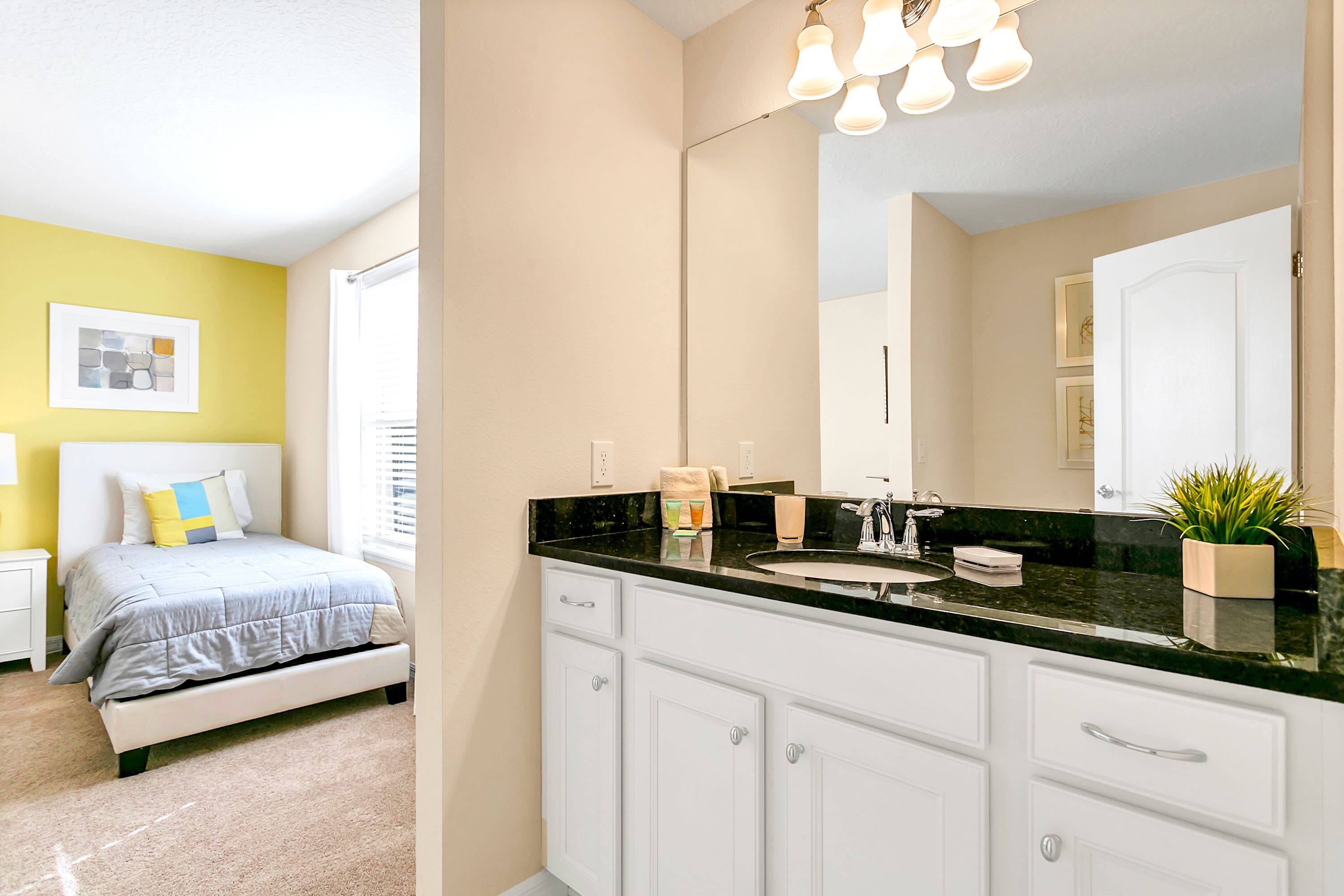 A bathroom vanity with a dark granite countertop, white cabinets, and a mirror reflecting a bedroom with a yellow accent wall and a bed.