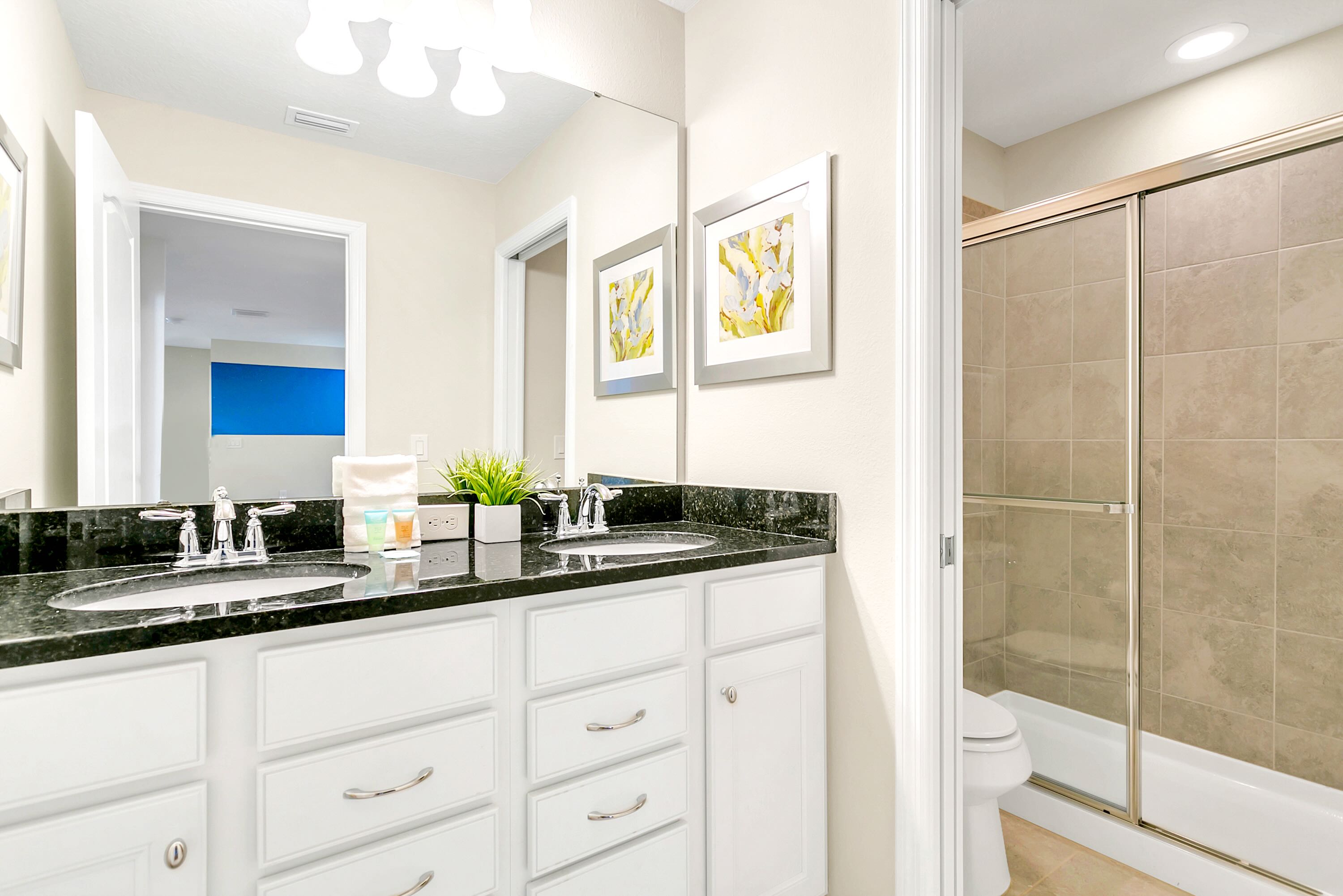 A bright bathroom with a double vanity, dark granite countertops, white cabinets, and a large mirror reflecting a bedroom with a yellow accent wall. The shower-tub combo with a sliding glass door and toilet are visible to the right.