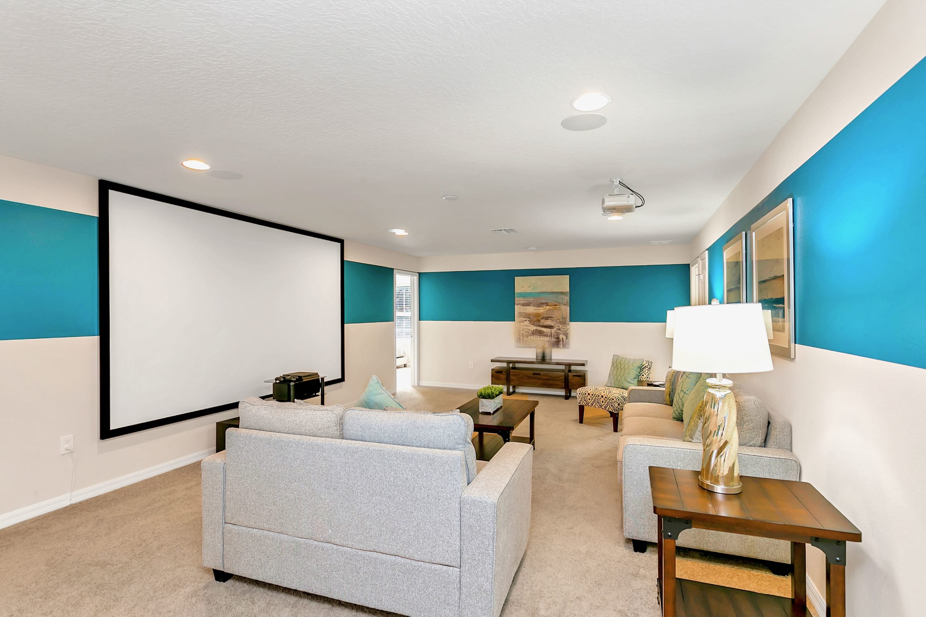 Dedicated media room featuring a large projection screen, two light gray sofas, and a patterned armchair. The walls are decorated with a striking horizontal blue stripe, and the floor is carpeted.
