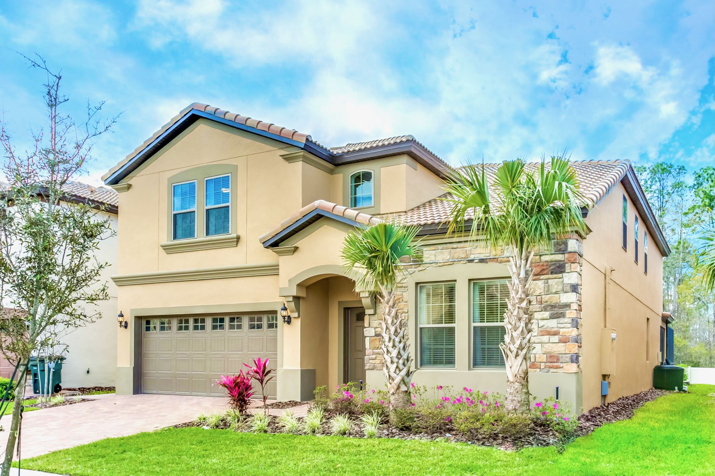 Two-story stucco house with a tiled roof, a two-car garage, and a stone facade accent, featuring a well-maintained green lawn and tropical landscaping with palm trees and flowering plants.