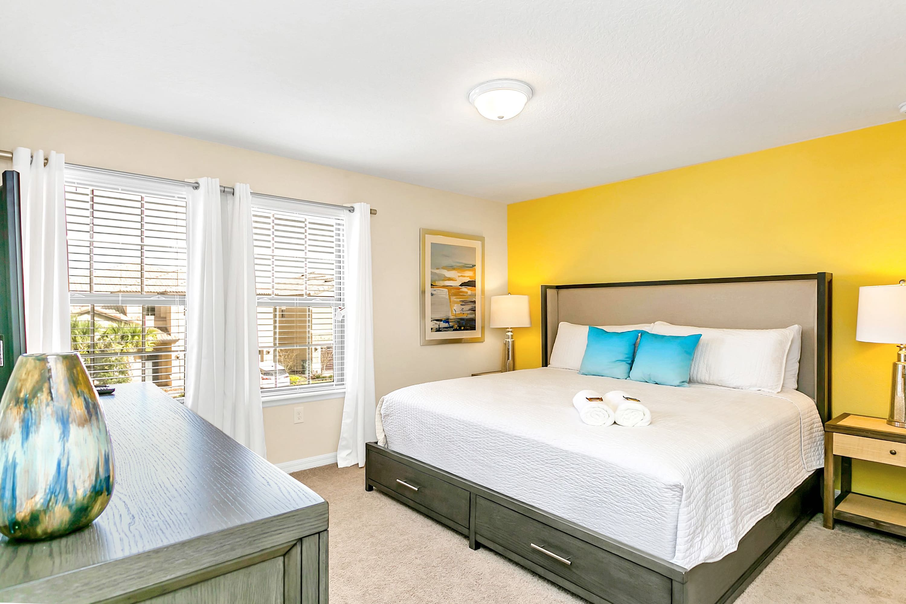 A spacious bedroom with a king-sized bed featuring a gray upholstered headboard, a vibrant yellow accent wall, and two large windows with white blinds and curtains. A dark gray dresser and a nightstand with a lamp are also present.