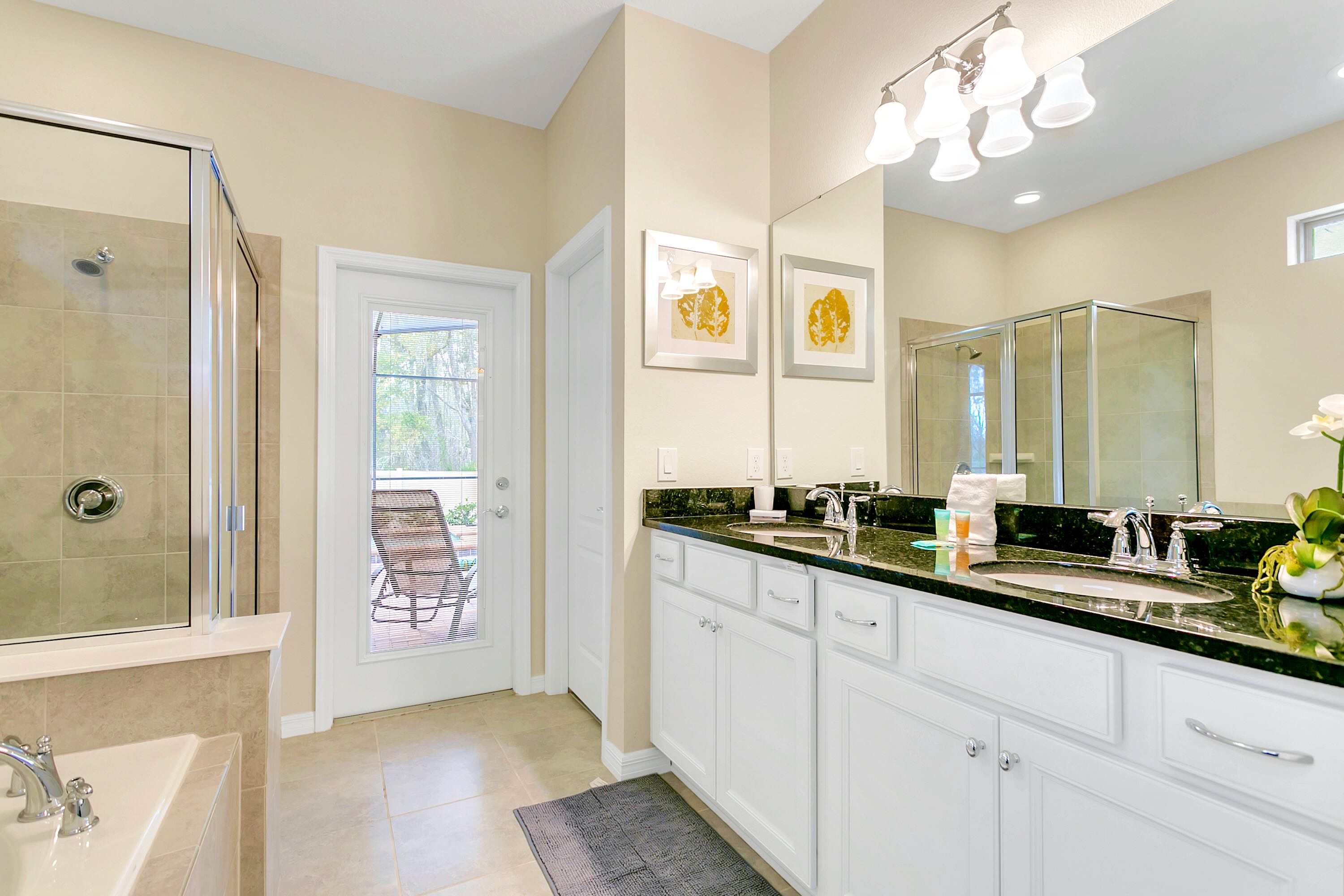 A bright bathroom featuring a double vanity with dark granite countertops, white cabinets, and two framed abstract artworks above the sinks. A glass-enclosed shower and a door leading to an outdoor patio are visible.