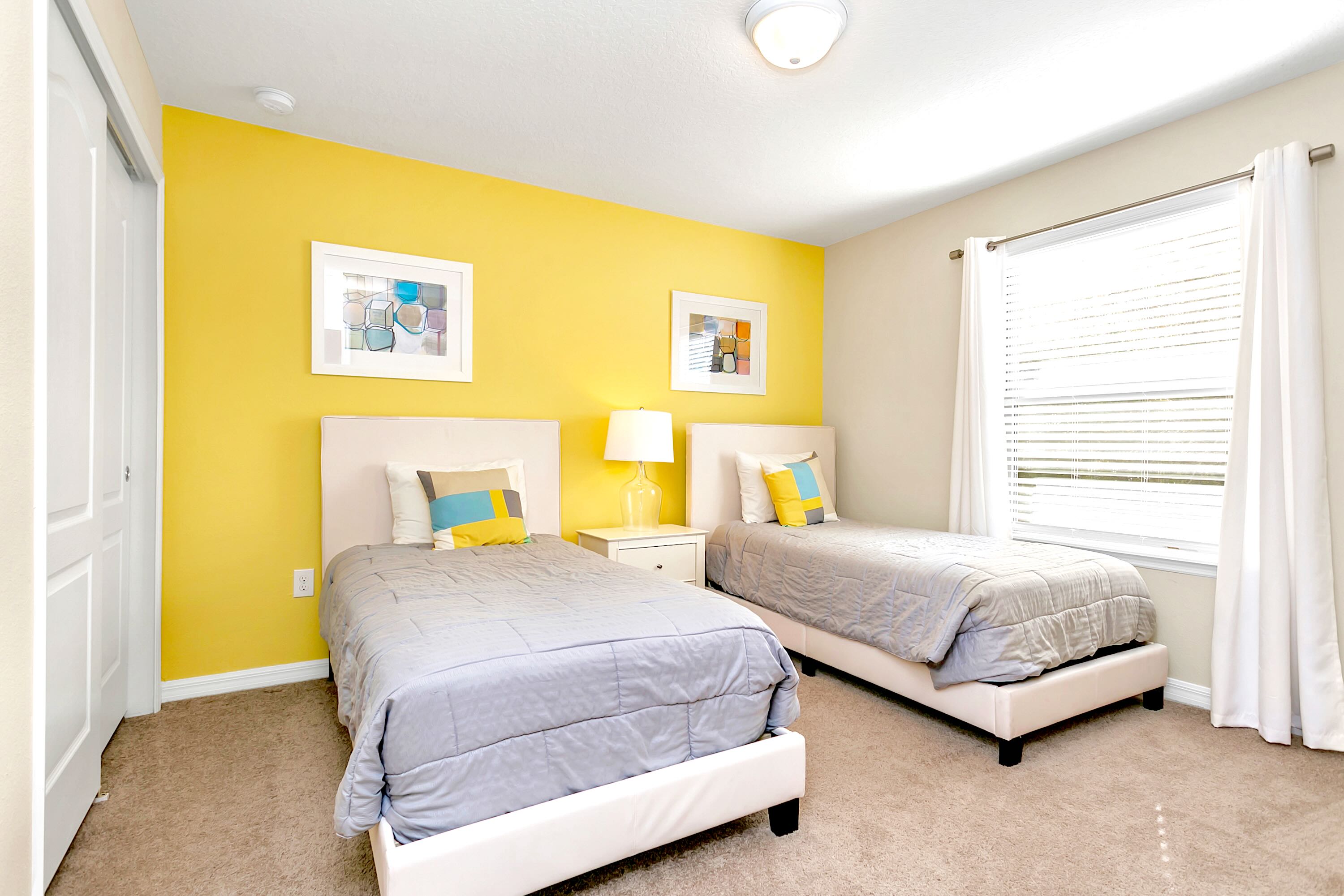 This bright bedroom features two twin beds with light-colored frames and grey bedding, a striking yellow accent wall, and a window with white blinds and curtains.