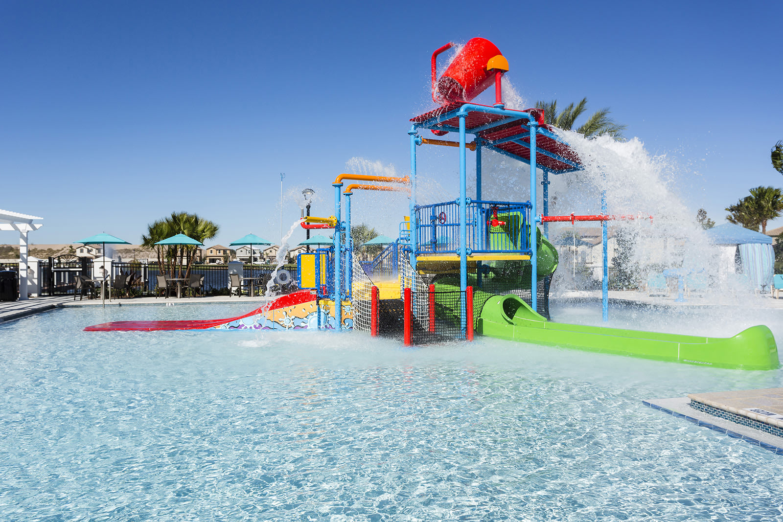 A vibrant children's splash pad area within a communal pool, featuring colorful slides, climbing structures, and a large red dumping bucket actively spilling water. The surrounding pool water is clear and blue under a bright sky.