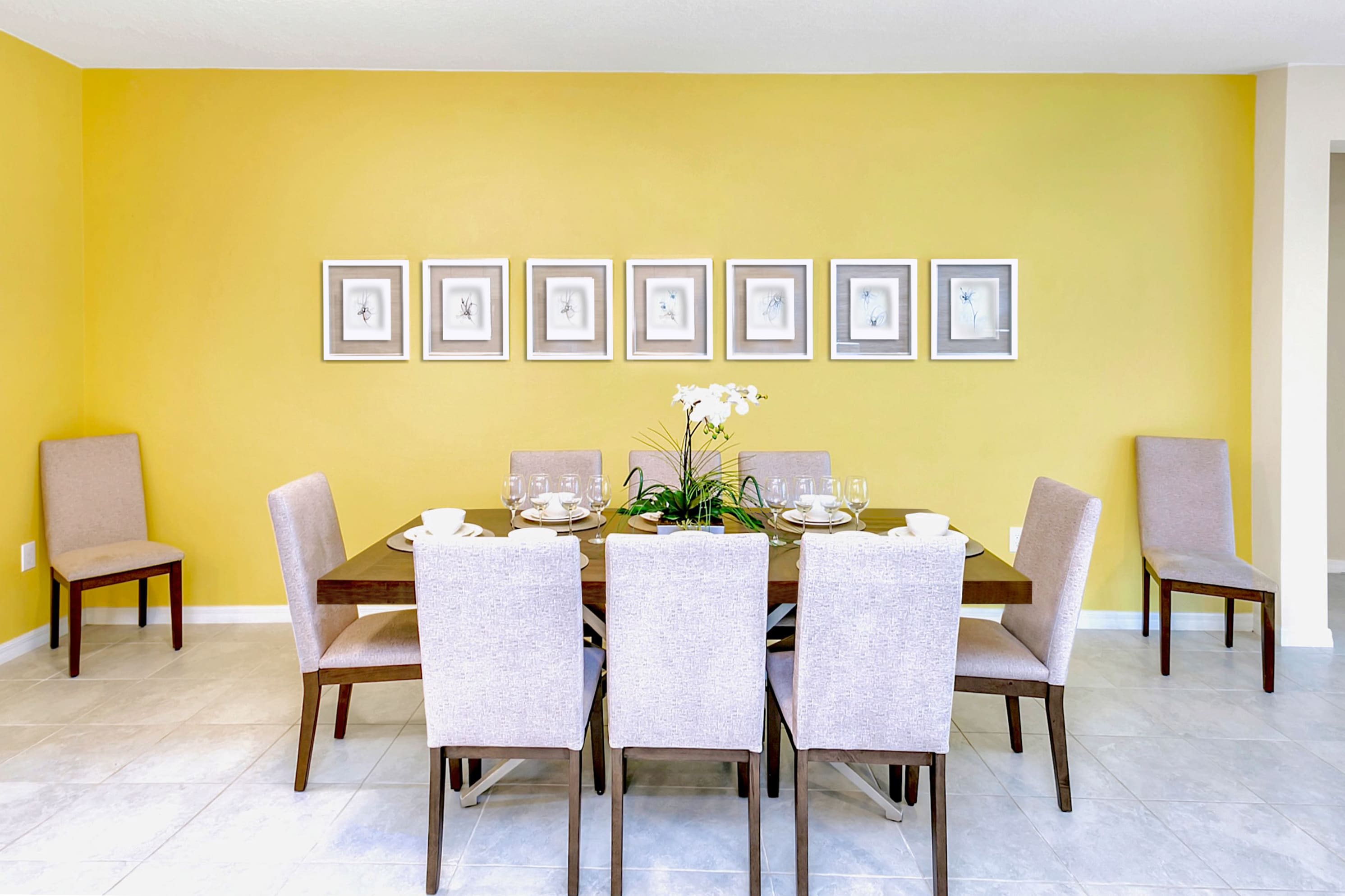 Elegant dining room featuring a long wooden table set for eight, surrounded by upholstered chairs. A vibrant yellow accent wall is adorned with a symmetrical row of eight framed botanical prints.