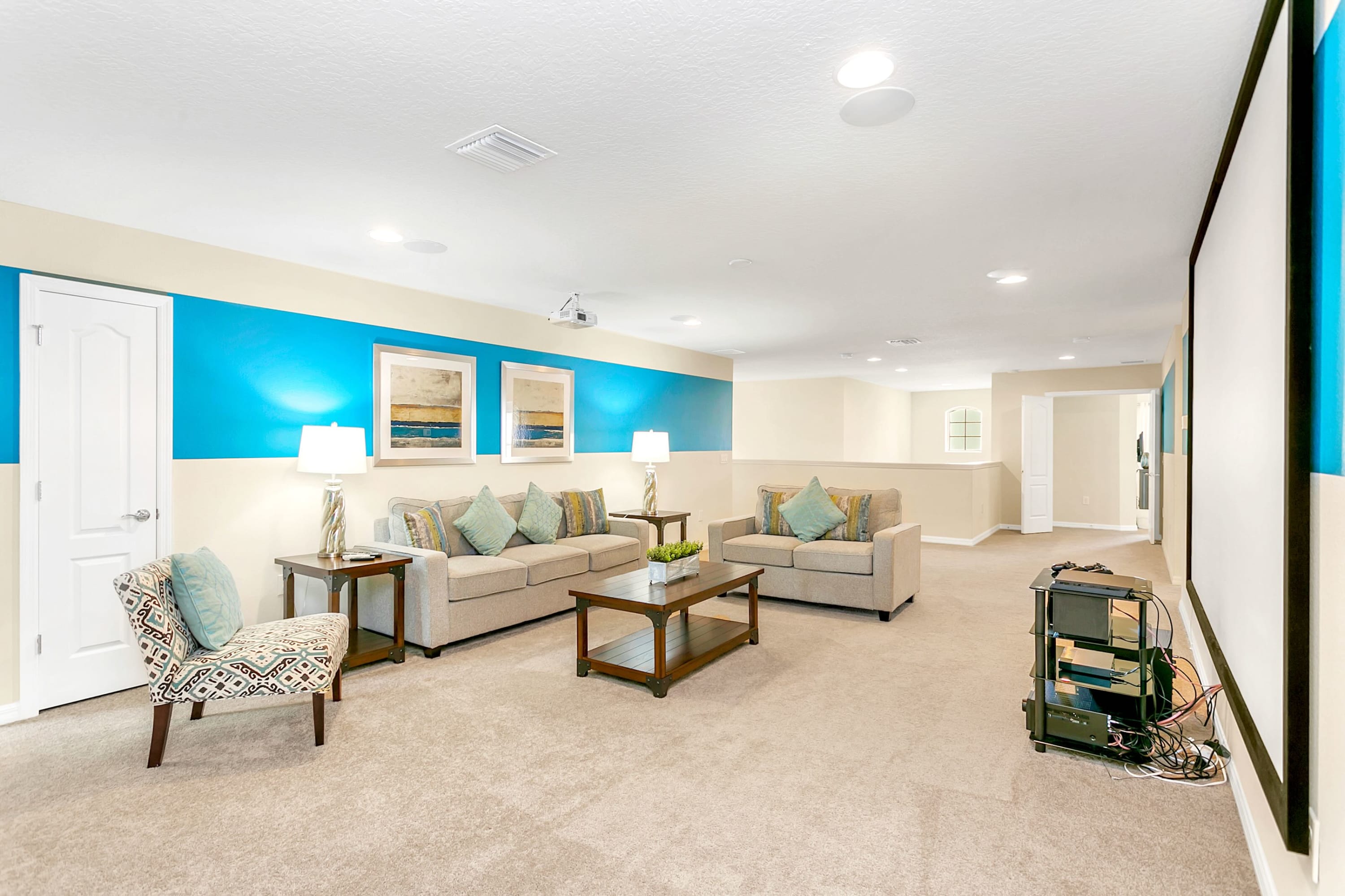 Spacious media room with two comfortable light gray sofas, a wooden coffee table, and side tables with lamps. The room features a bold blue stripe on the walls and soft beige carpeting.