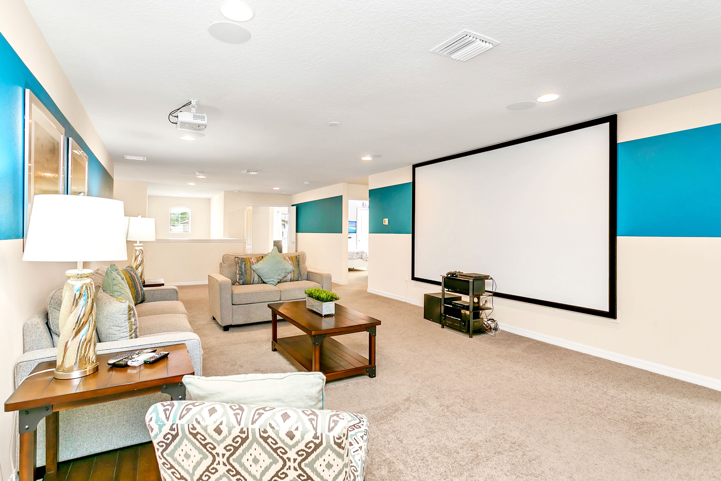 Spacious media room featuring a large projection screen, comfortable sofas, and armchairs, with a coffee table, carpeted floor, and accent walls with horizontal blue stripes.