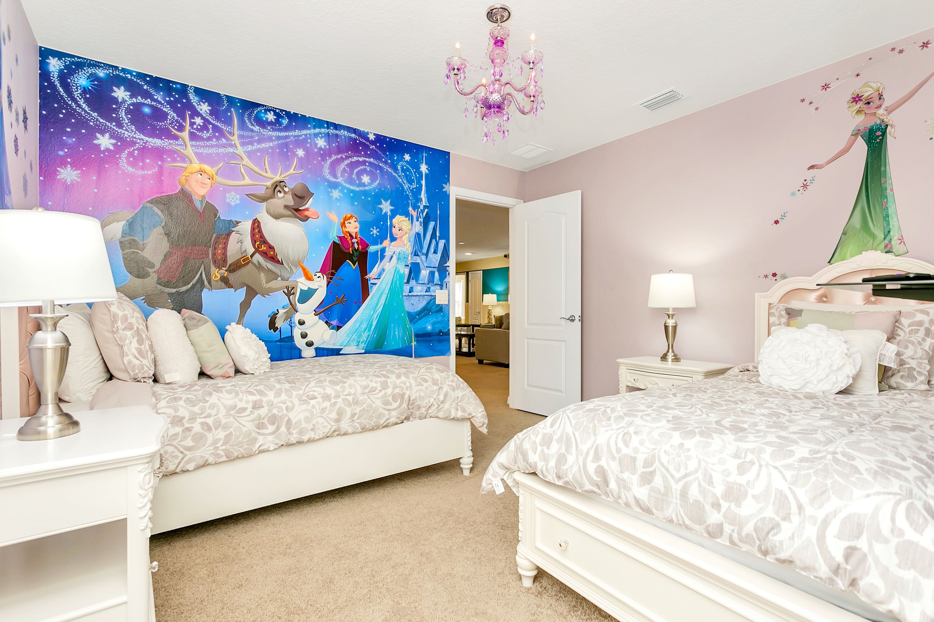A vibrant kids' bedroom features two twin beds with white frames and patterned bedding, a large "Frozen" mural on one wall, and a pink chandelier.