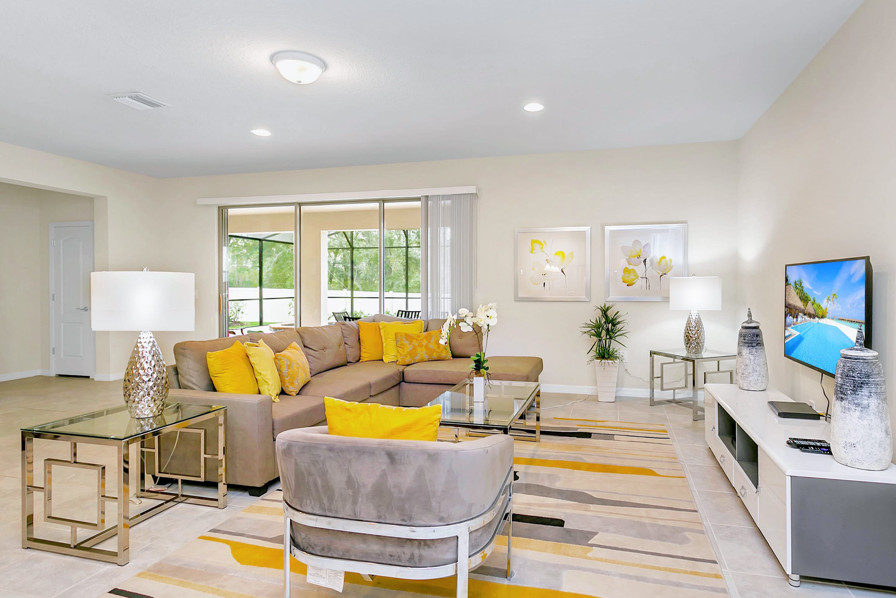 Modern living room with a large gray sectional sofa, an armchair, a glass coffee table, and a striped rug, featuring a wall-mounted TV on a media console and sliding glass doors leading to an outdoor area.