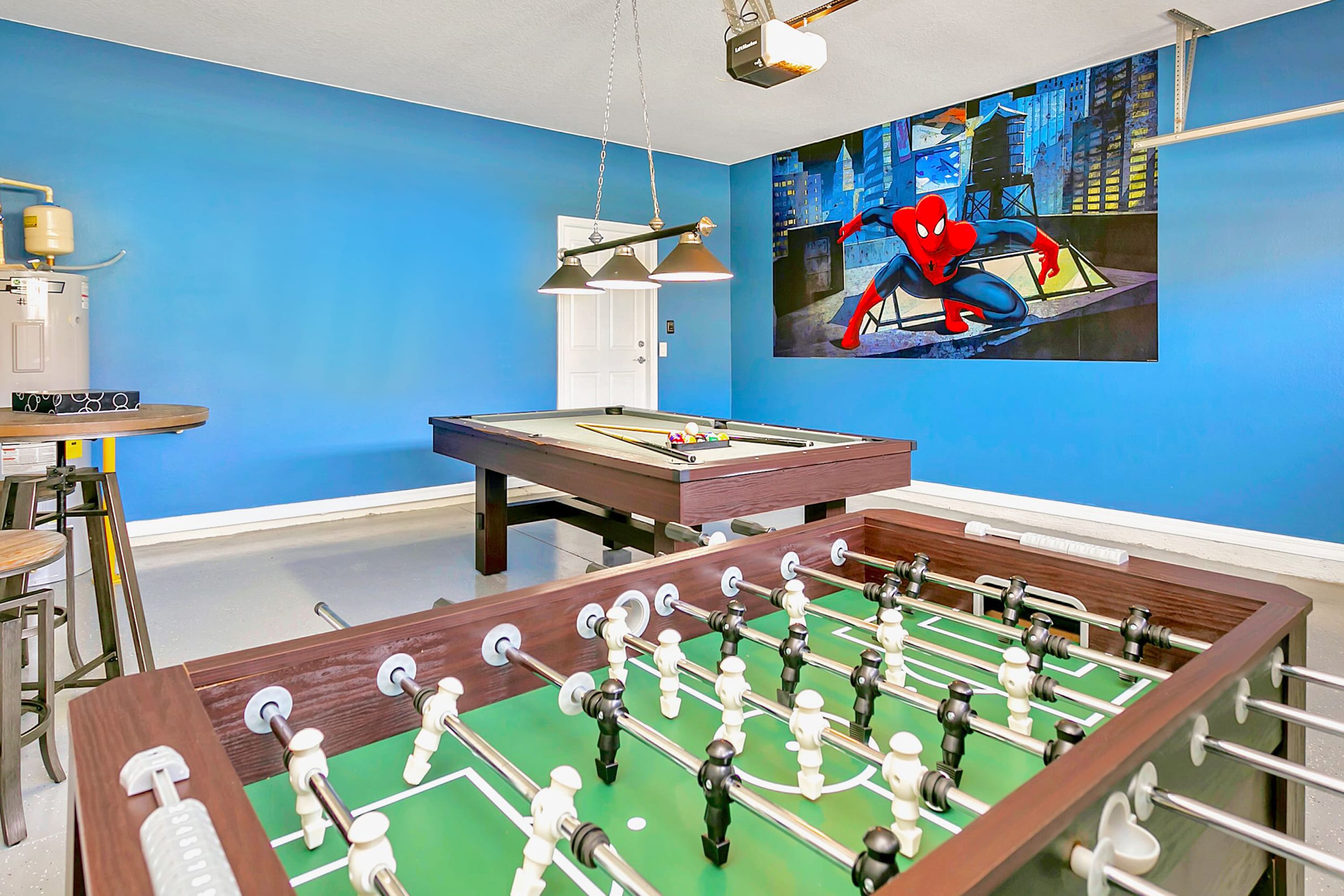 Vibrant game room in a garage setting, featuring a pool table and a foosball table, with bright blue walls and a large Spiderman mural, illuminated by industrial pendant lights.