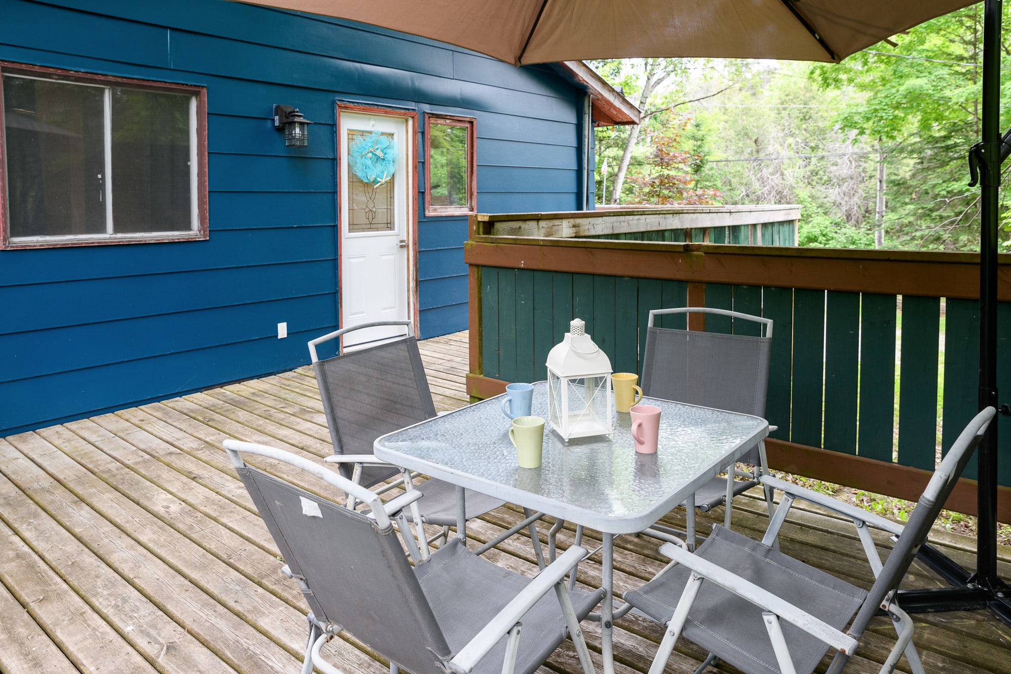 A section of a wooden deck featuring a glass-top outdoor dining table with four gray folding chairs, shaded by a large brown umbrella. A blue house exterior is visible in the background, surrounded by green trees.