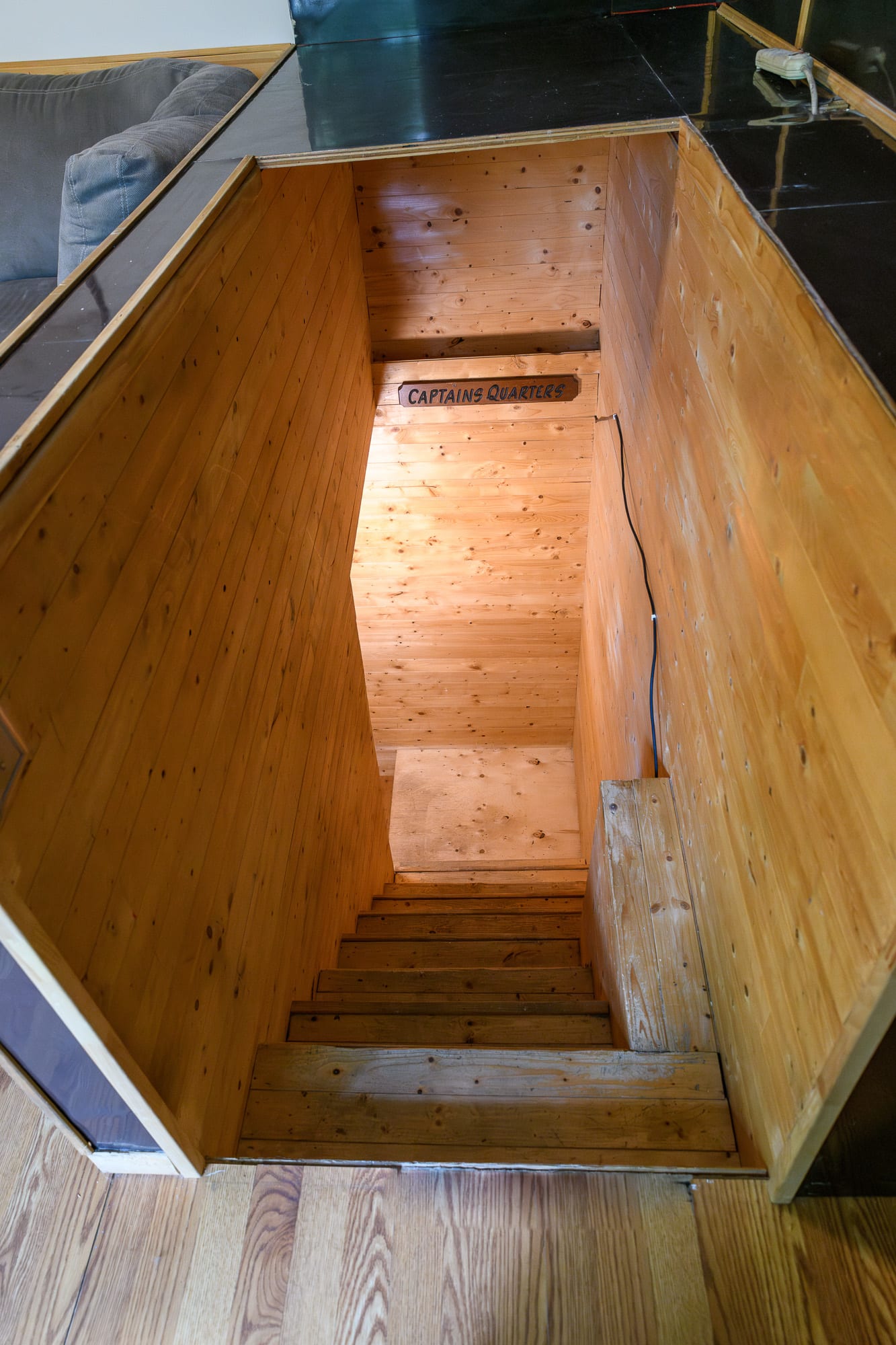 A narrow wooden staircase leading downwards, with a sign above reading 'CAPTAINS QUARTERS'. The walls and steps are constructed from light-colored wood planks, creating a rustic feel.
