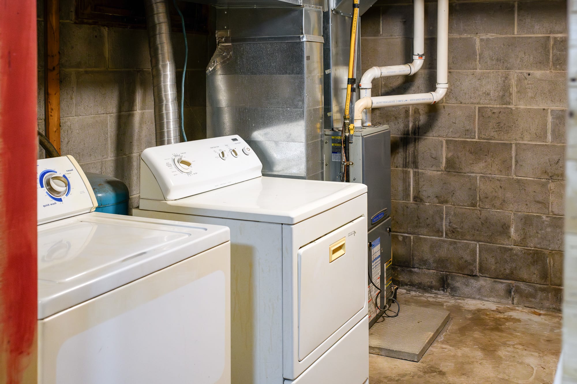 A functional laundry area with a white top-loading washing machine and a white dryer, positioned against a gray cinder block wall. Exposed ductwork and pipes are visible above the appliances, indicating a utility space.