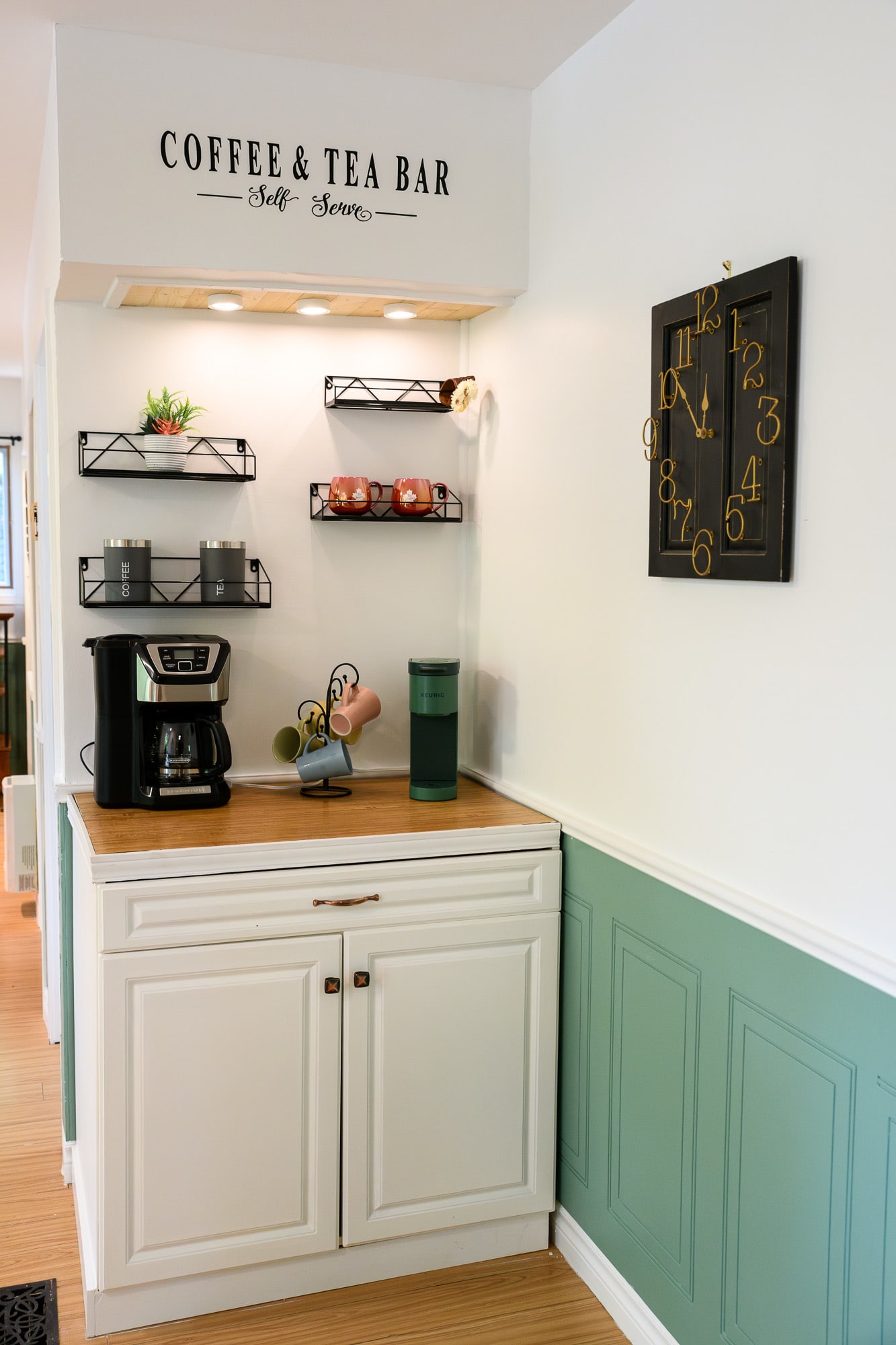 A dedicated coffee and tea bar area with a white cabinet base, light wood-look countertop, coffee maker, and wall-mounted shelves displaying mugs. A 'COFFEE & TEA BAR' sign is above, and a black decorative clock hangs on the wall.
