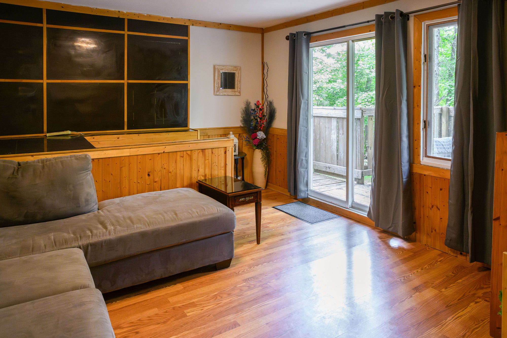A bright living room with light wood-look flooring and a gray sectional sofa, featuring a large sliding glass door leading to an outdoor deck. The room has wood paneling along the lower walls and dark gray curtains.