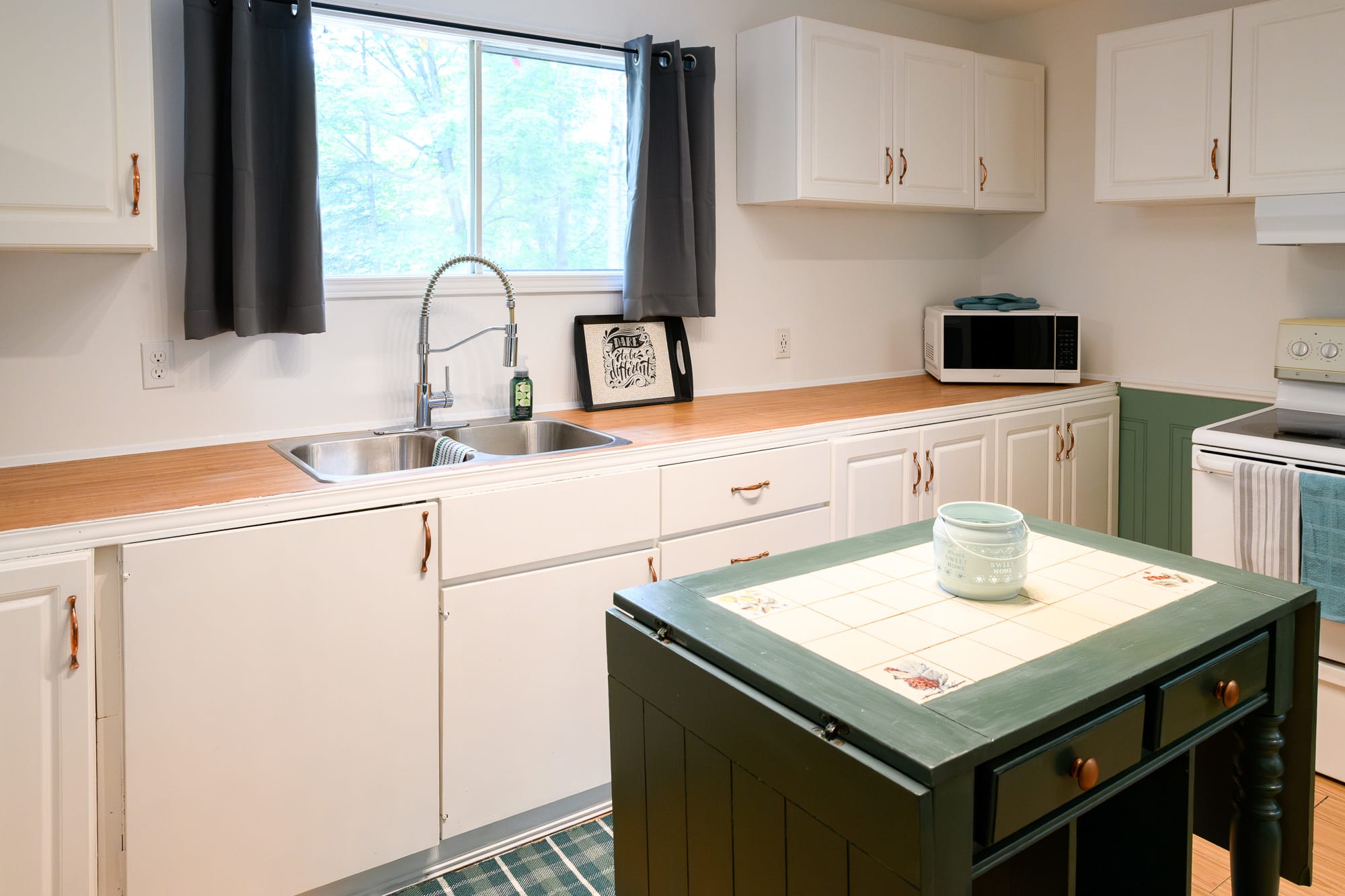 A bright kitchen featuring white shaker-style cabinets, light wood-look countertops, a double stainless steel sink, and a central dark green island with a tiled top. A microwave sits on the counter, and a window overlooks trees.
