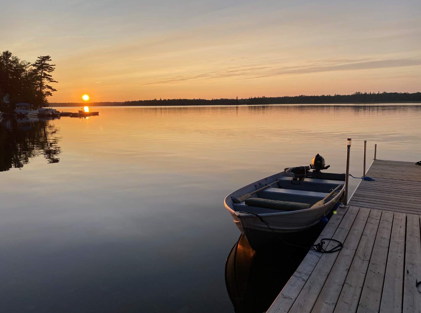 A tranquil sunset over a calm lake, featuring a wooden dock extending into the water with a small boat tied alongside. The sky is painted with warm orange and yellow hues, reflecting softly on the water's surface.