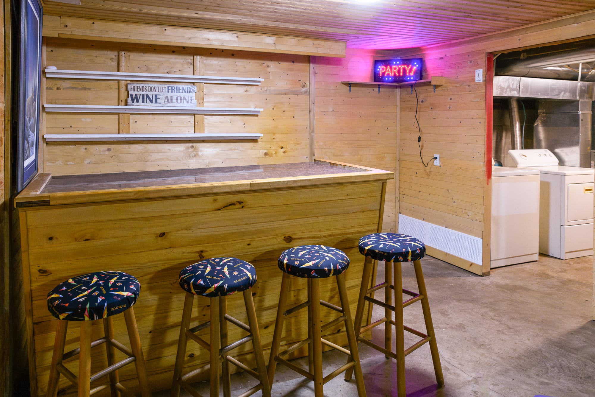 A rustic wooden bar with four stools featuring colorful patterned cushions, illuminated by a "PARTY!" neon sign above. The bar is set against wood-paneled walls, with a laundry area including a washer and dryer visible through an opening in the background.