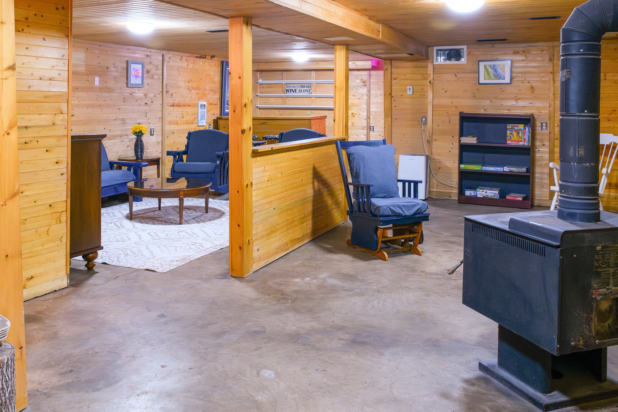 A rustic basement living area with wood-paneled walls and ceiling, featuring a blue sofa and armchairs on a patterned rug, a round wooden coffee table, and a black wood-burning stove in the foreground. A wooden bar with stools is visible in the background.
