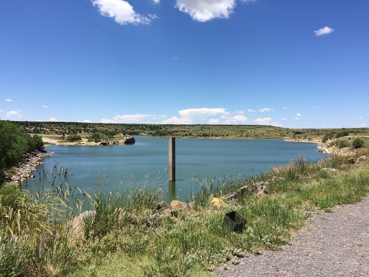 A wide shot of a serene lake or reservoir under a bright blue sky with scattered white clouds, surrounded by rolling hills covered in sparse green vegetation. A tall, dark pillar stands in the water, and a gravel path runs along the foreground.