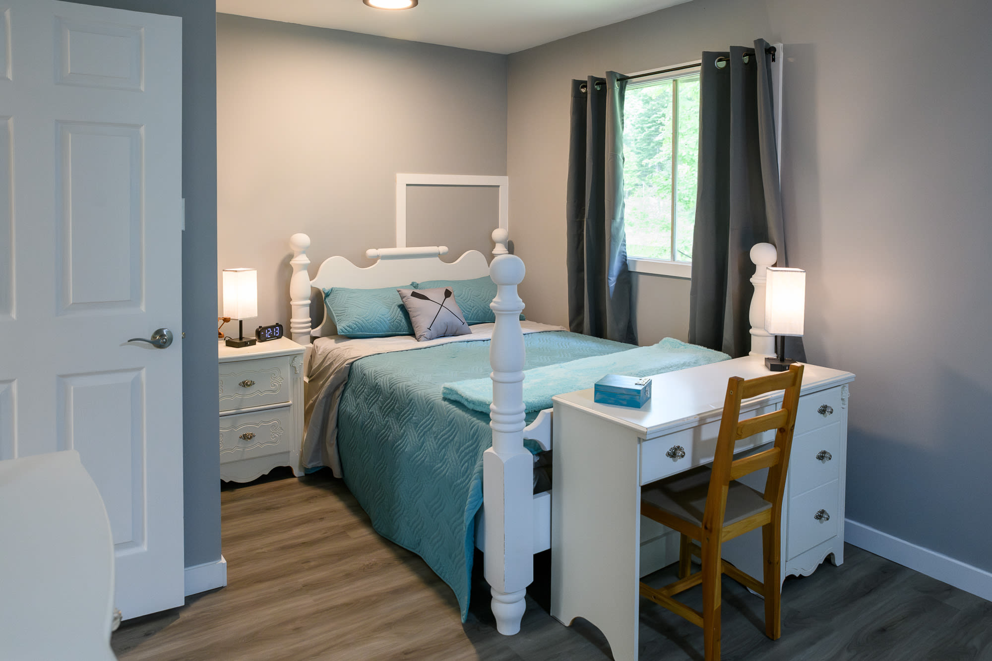 A bedroom featuring a white full-sized bed with a blue comforter and pillows, complemented by a white desk and chair. White nightstands with lamps flank the bed, and a window provides natural light.