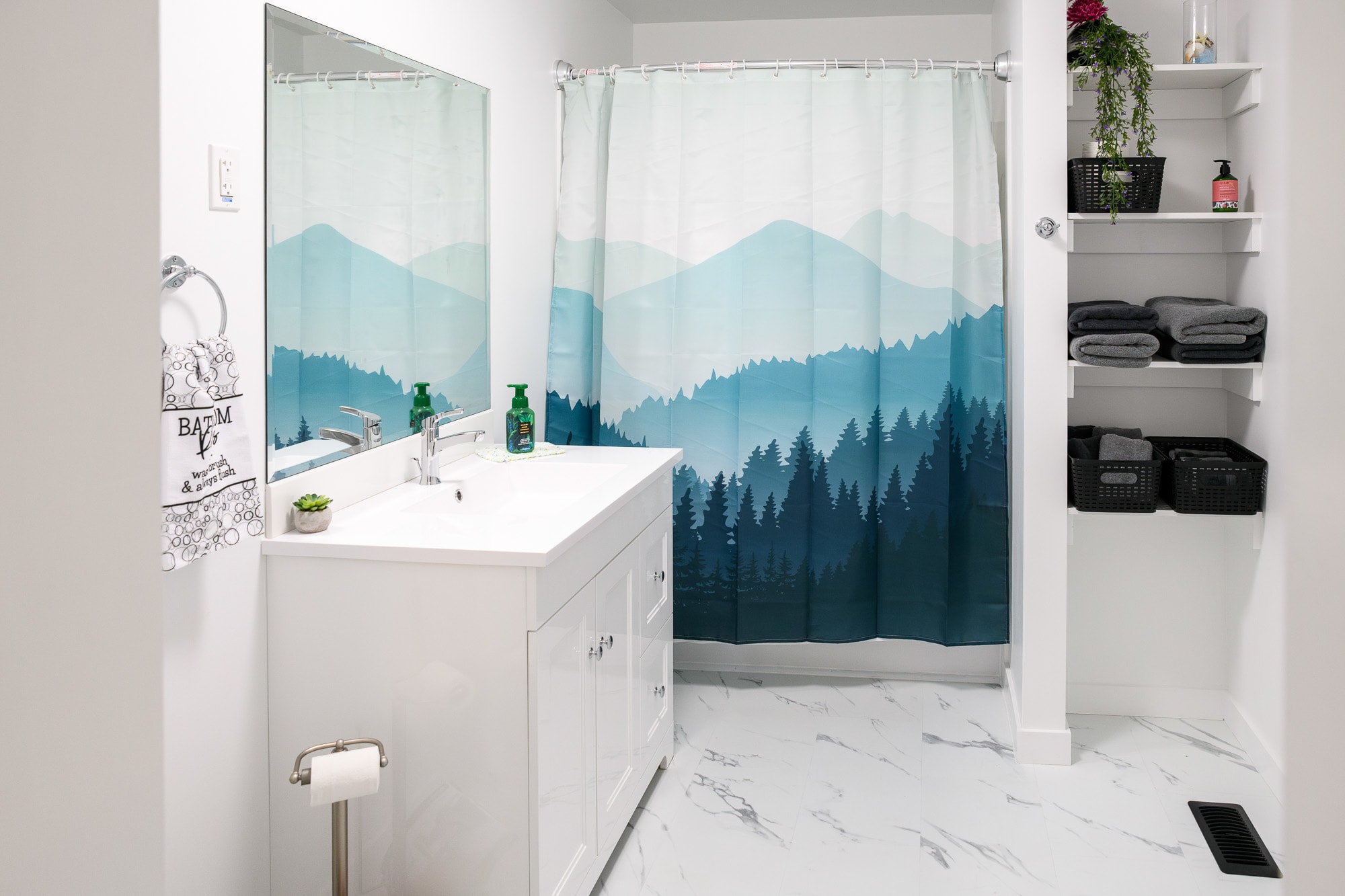 A bright bathroom with a white vanity, large mirror, and a shower/tub combination enclosed by a blue and green mountain-themed shower curtain. Open shelving stocked with towels and baskets is built into the wall on the right.