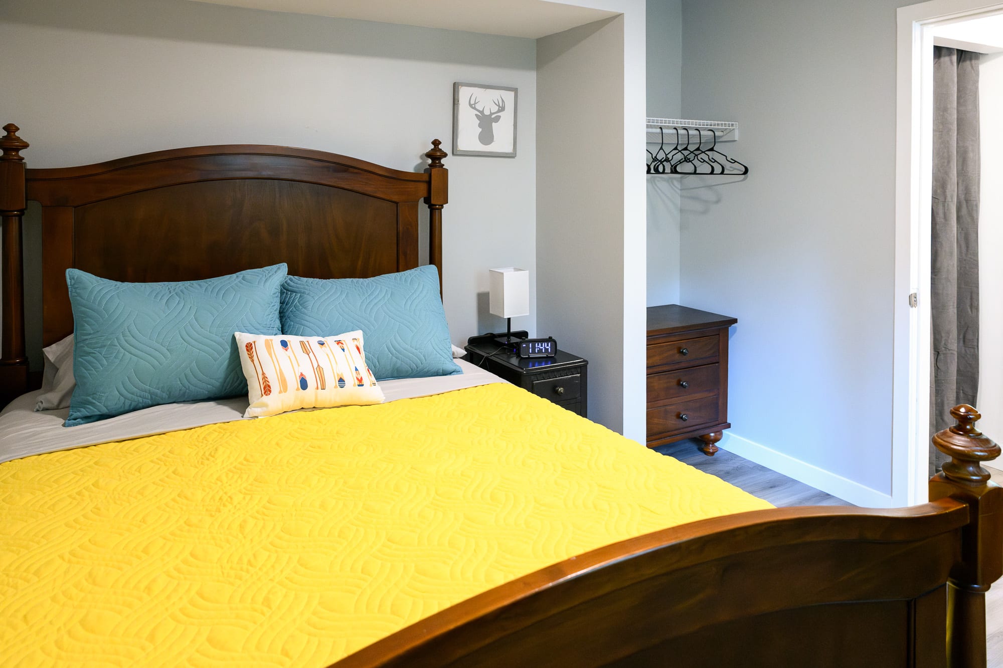 A cozy bedroom featuring a dark wood queen-sized bed with a tall, ornate headboard, dressed in a bright yellow quilted comforter and blue and white throw pillows. A small dark nightstand with a lamp sits beside the bed.
