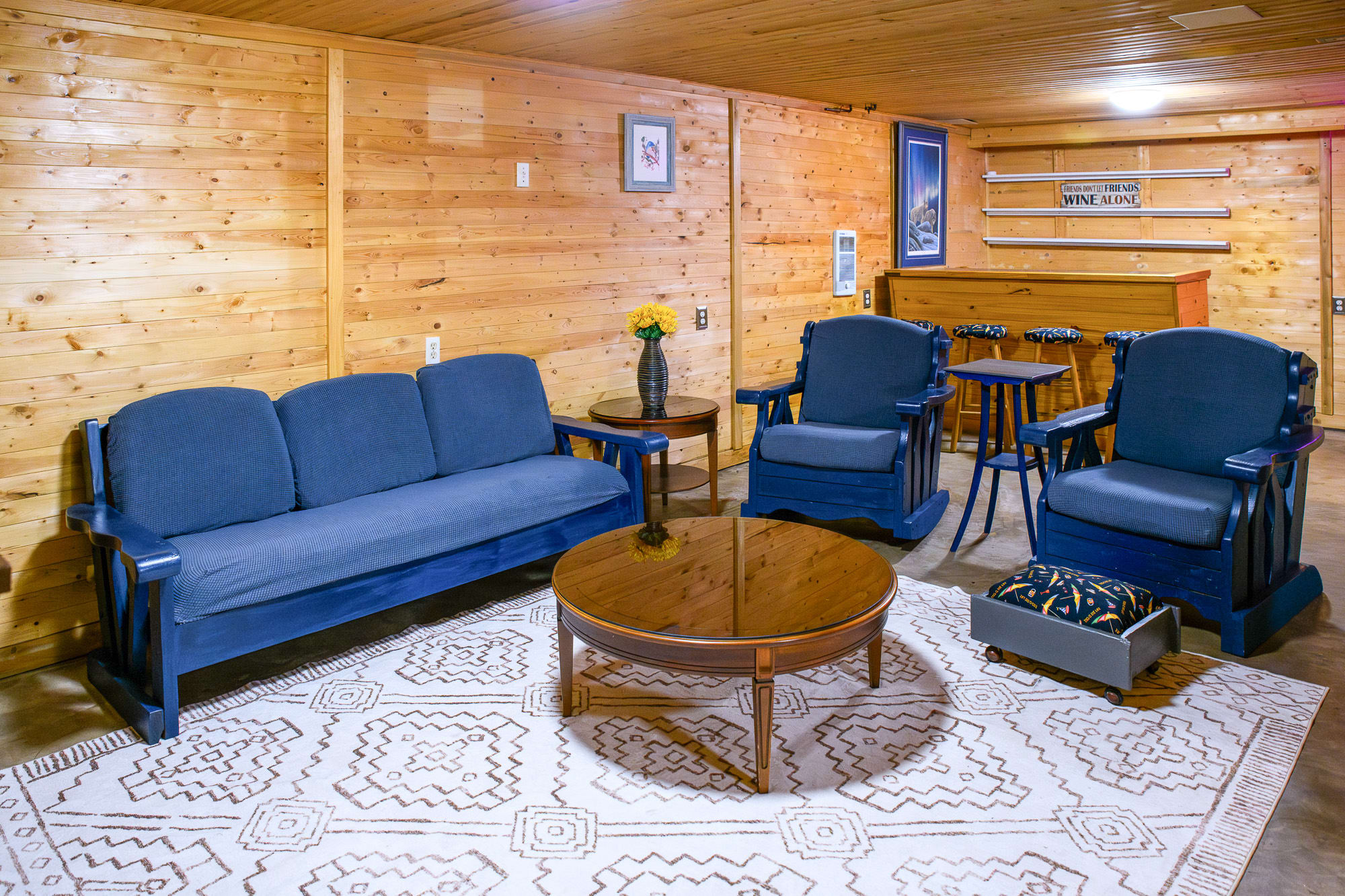 A rustic living area with warm wood-paneled walls, furnished with a blue sofa, two blue armchairs, and a round wooden coffee table centered on a patterned area rug. A bar area is visible in the background.
