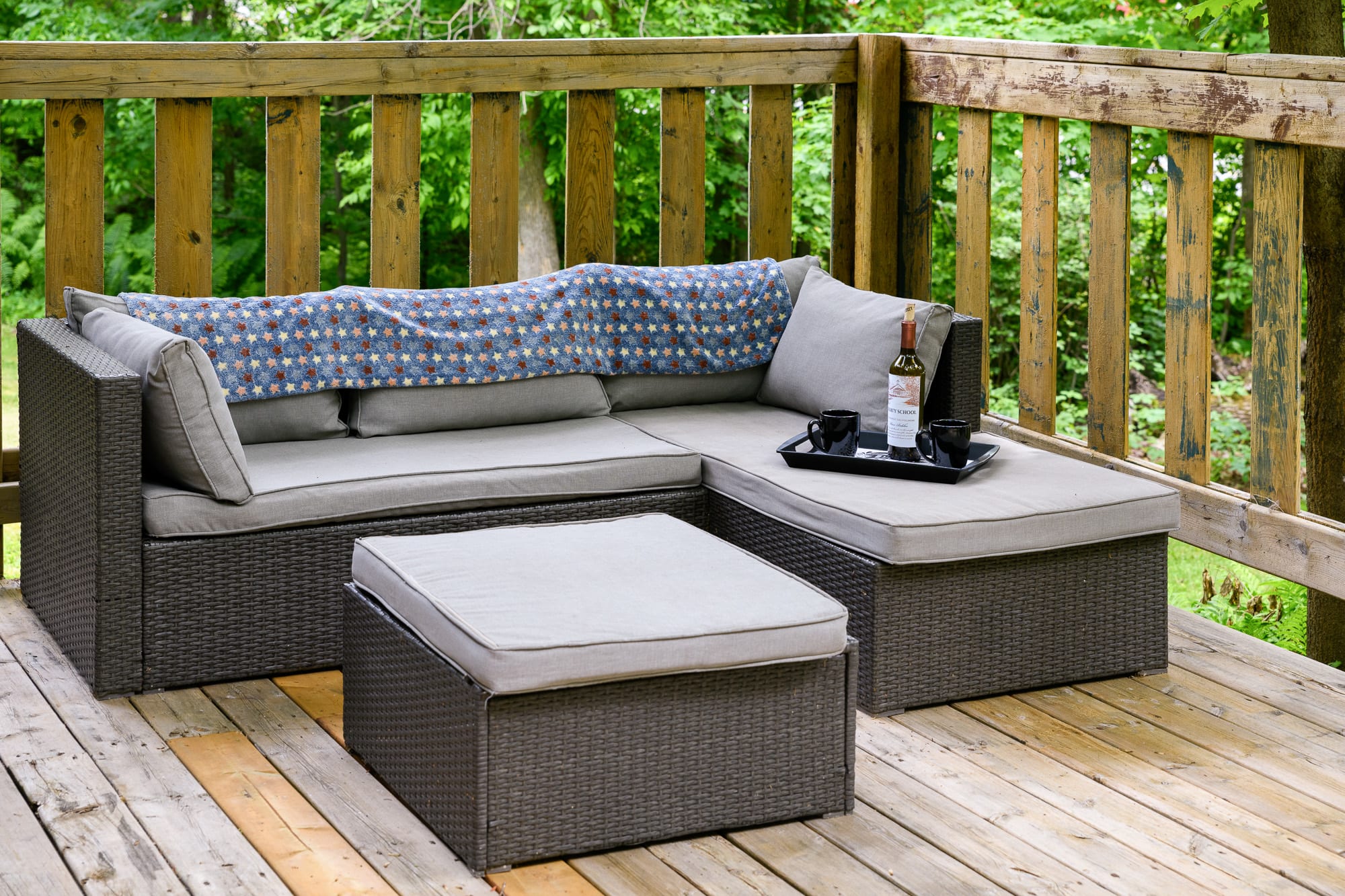 A cozy section of a wooden deck with a dark green railing, furnished with a gray wicker sectional sofa and an ottoman, adorned with a patterned blanket and a tray with a bottle and cups. Lush green trees are visible in the background.