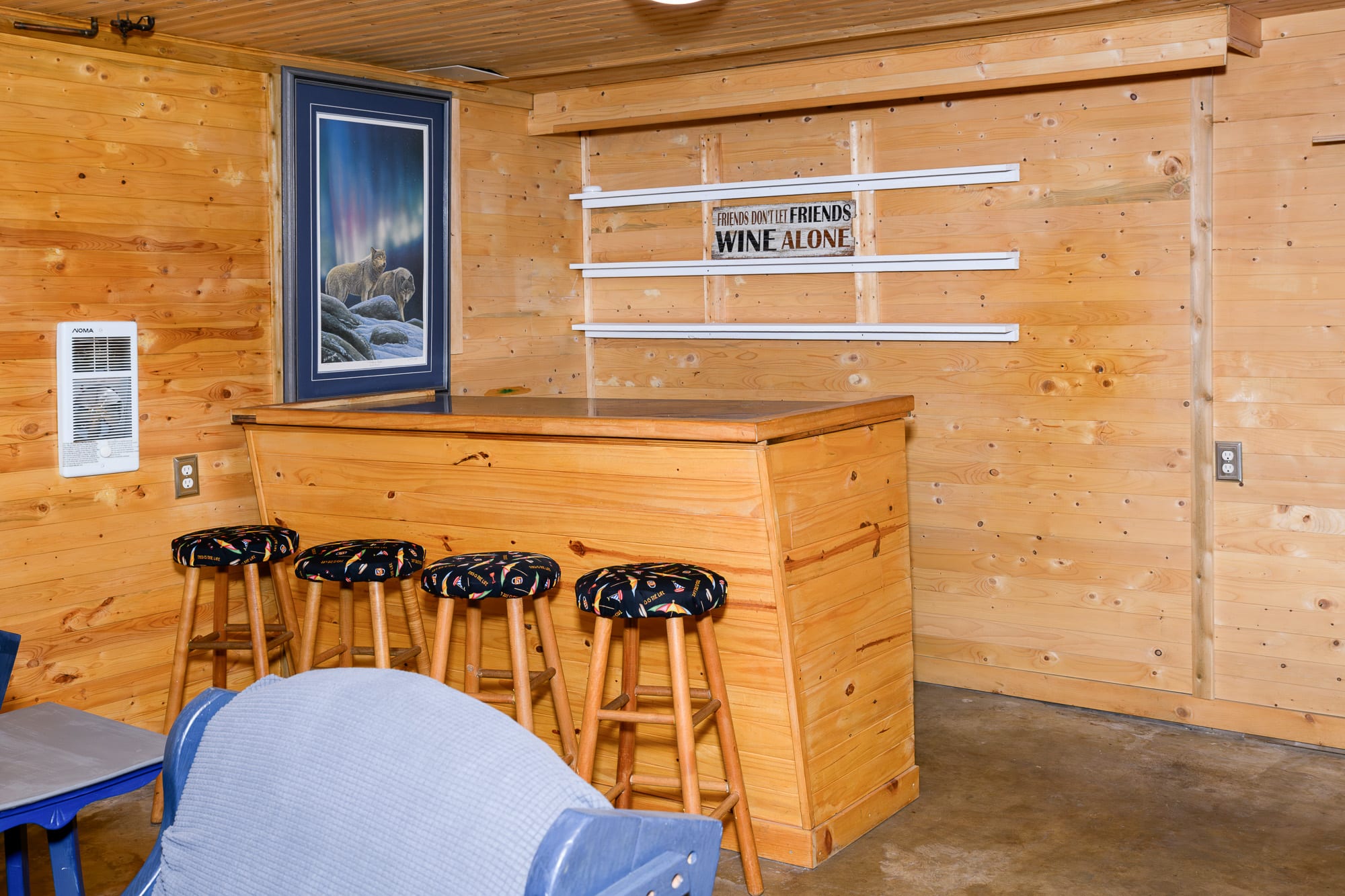 A wooden bar with a dark countertop and three bar stools with patterned cushions, set against a light wood-paneled wall. A framed picture of wolves hangs above the bar, and a sign reading "FRIENDS DON'T LET FRIENDS WINE ALONE" is mounted on the wall above shelves.
