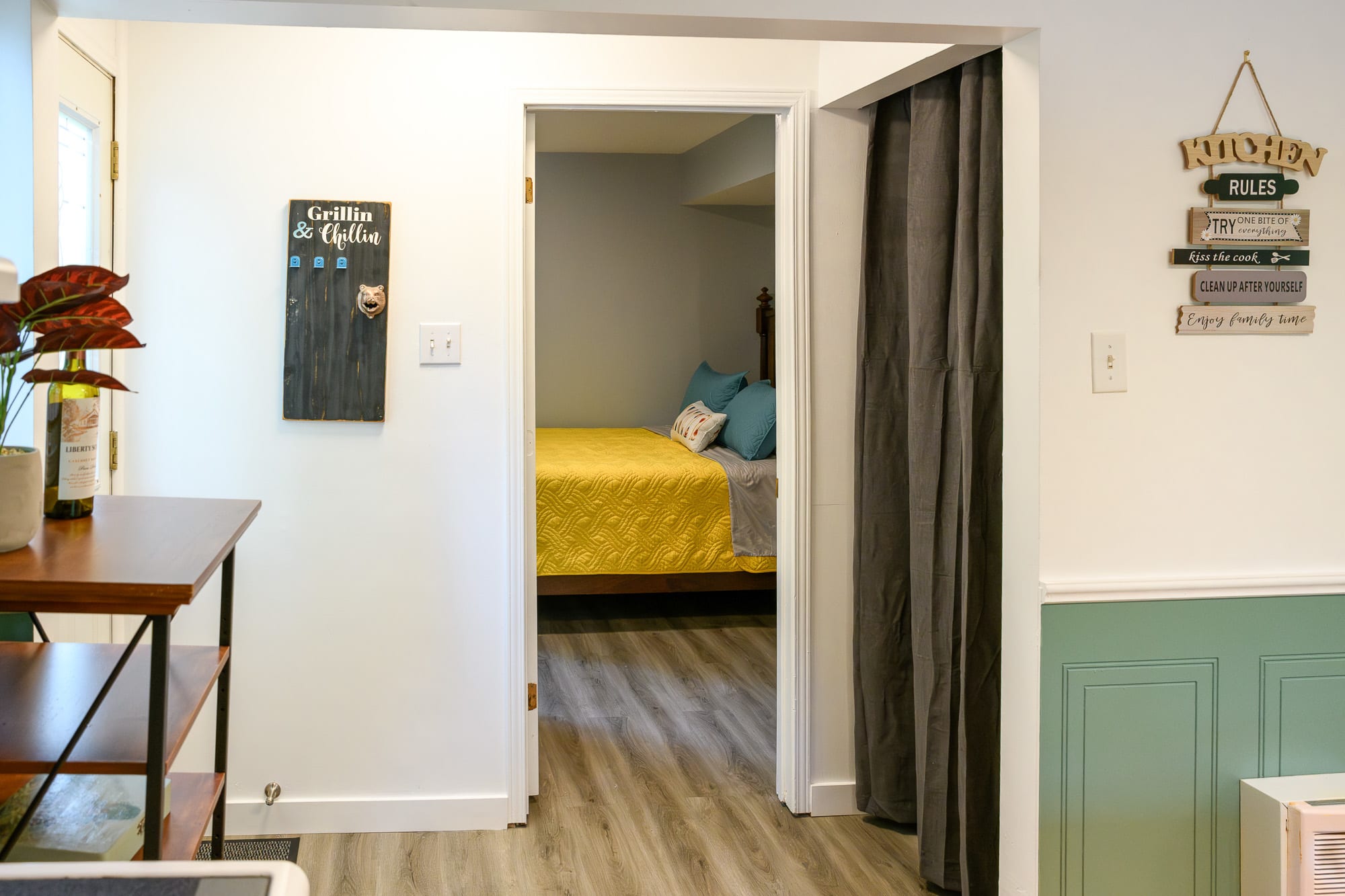 A hallway with white walls and green wainscoting, leading to a bedroom with a yellow bed visible through an open doorway. Decorative signs, including 'Grillin & Chillin' and 'KITCHEN RULES', adorn the walls.