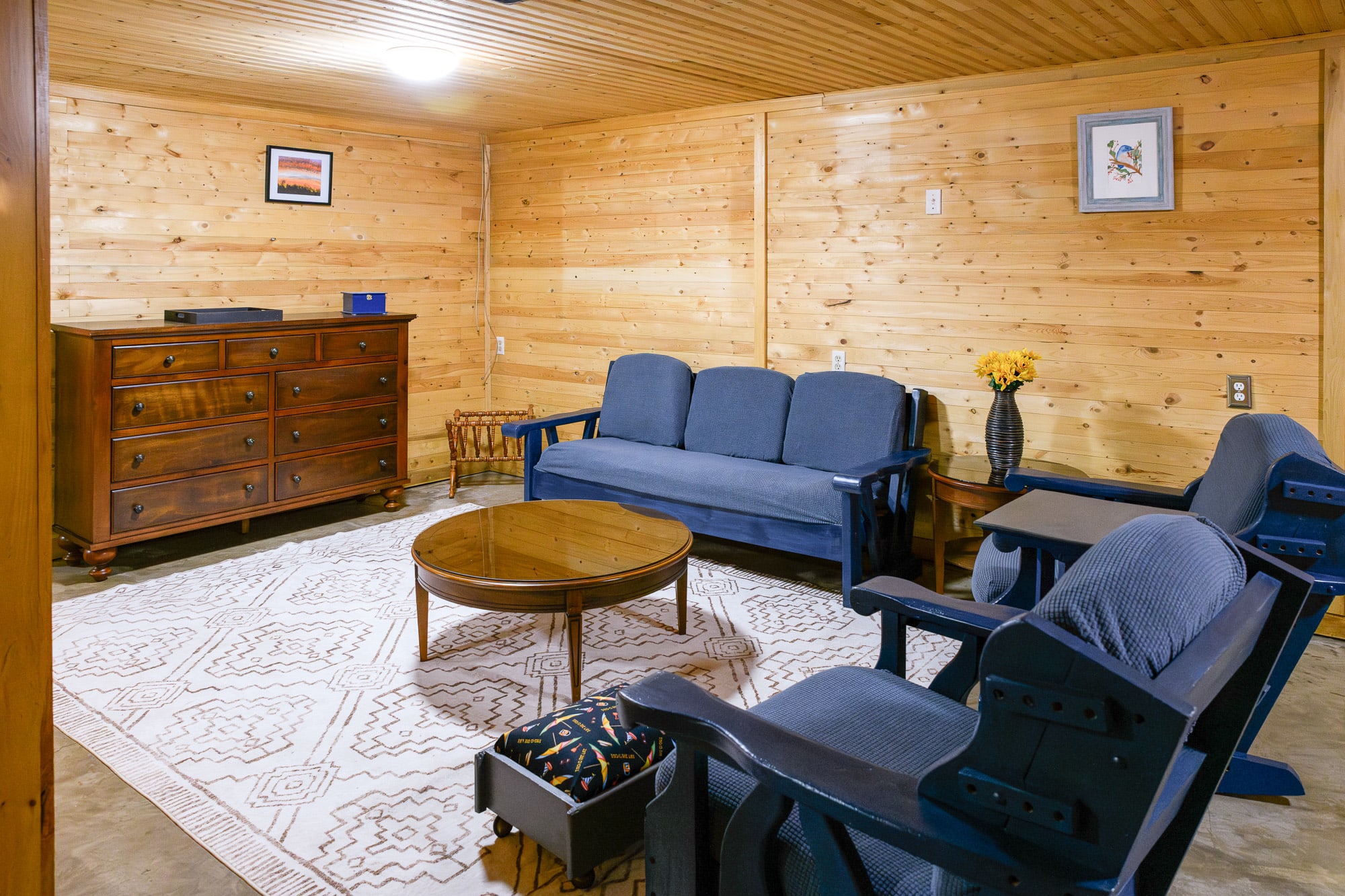 A cozy basement living space with light wood-paneled walls and ceiling, furnished with a blue three-seater sofa, two matching armchairs, and a round wooden coffee table centered on a large white patterned rug. A dark wooden dresser with a blue box and a vase of yellow flowers sits against the far wall.