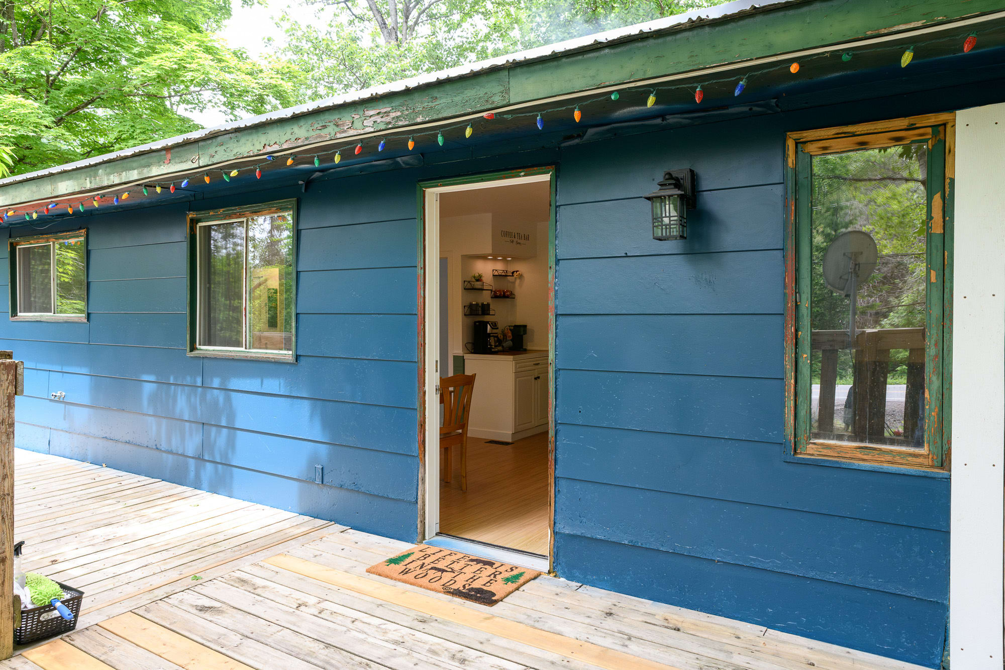 The vibrant blue exterior of a house with a white door leading to a wooden deck, adorned with colorful string lights along the roofline. Large windows reflect the surrounding greenery, and a "Welcome" mat is placed at the doorway.