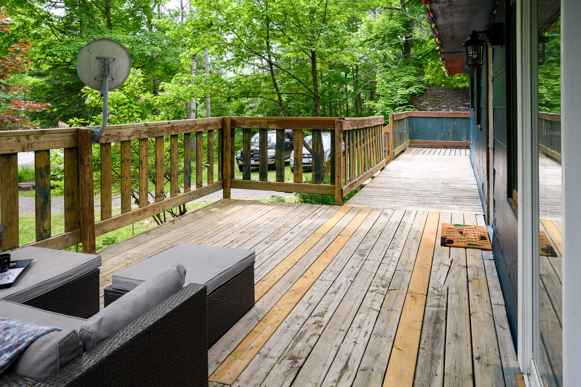 A spacious wooden deck with a natural wood railing, overlooking a lush green forest. Outdoor wicker seating with gray cushions is visible in the foreground, and a satellite dish is mounted on a post.