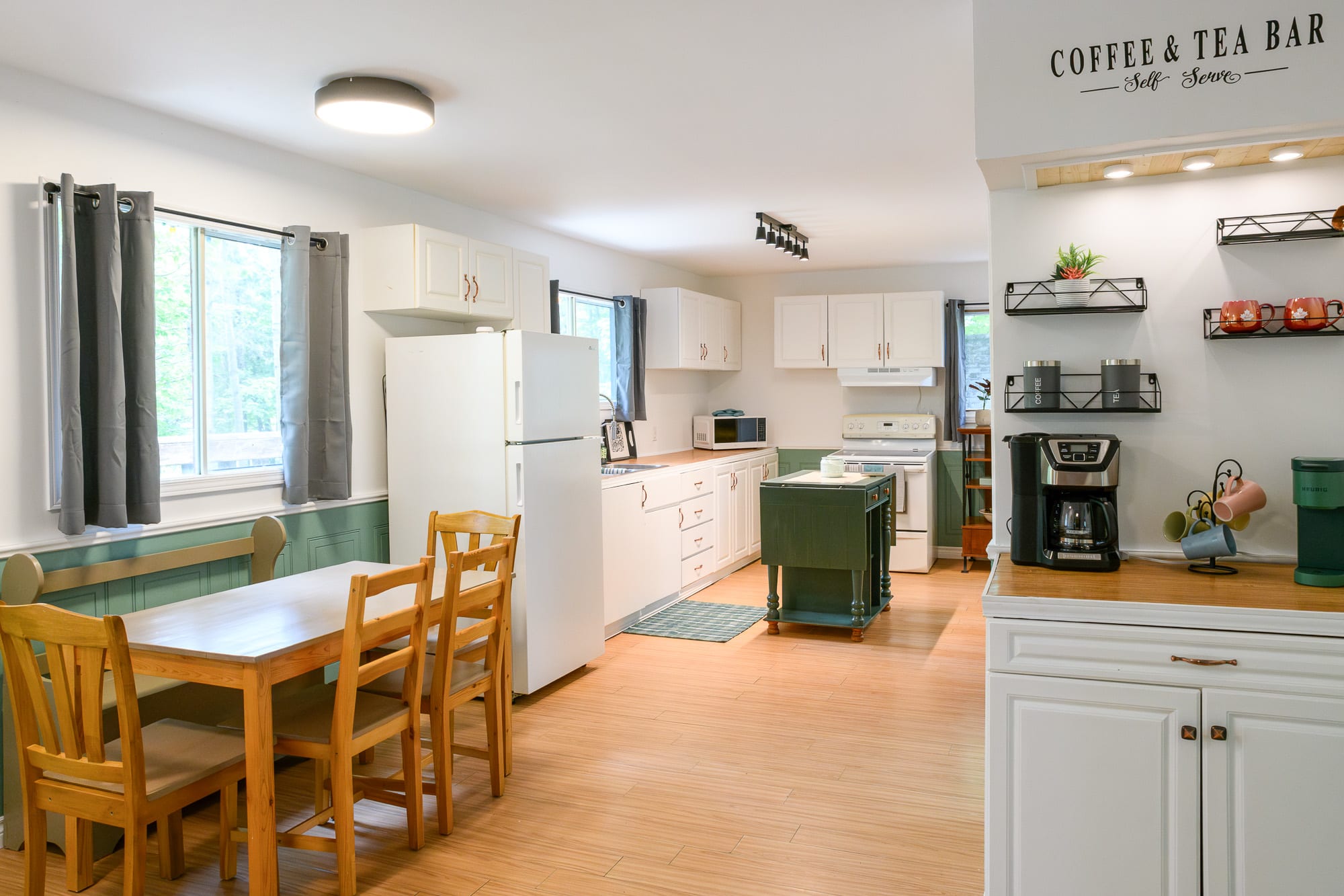 An open-concept kitchen and dining area with light wood-look flooring, white cabinets, and a "Coffee & Tea Bar" nook. The kitchen features a white refrigerator, stove, and microwave, while the dining area has a wooden table and chairs.