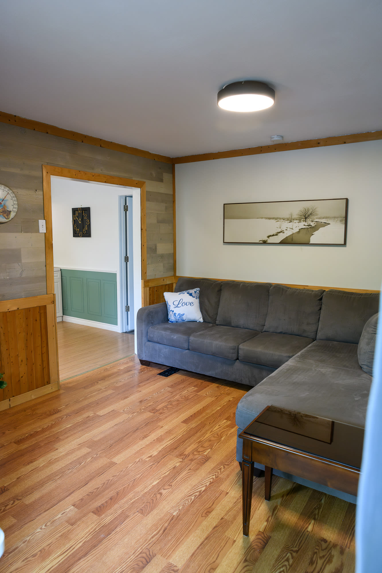 A cozy living room with light wood-look flooring and a gray sectional sofa, featuring wood paneling and a view into an adjacent room. A large landscape artwork hangs above the sofa, and a decorative clock is on the wall.